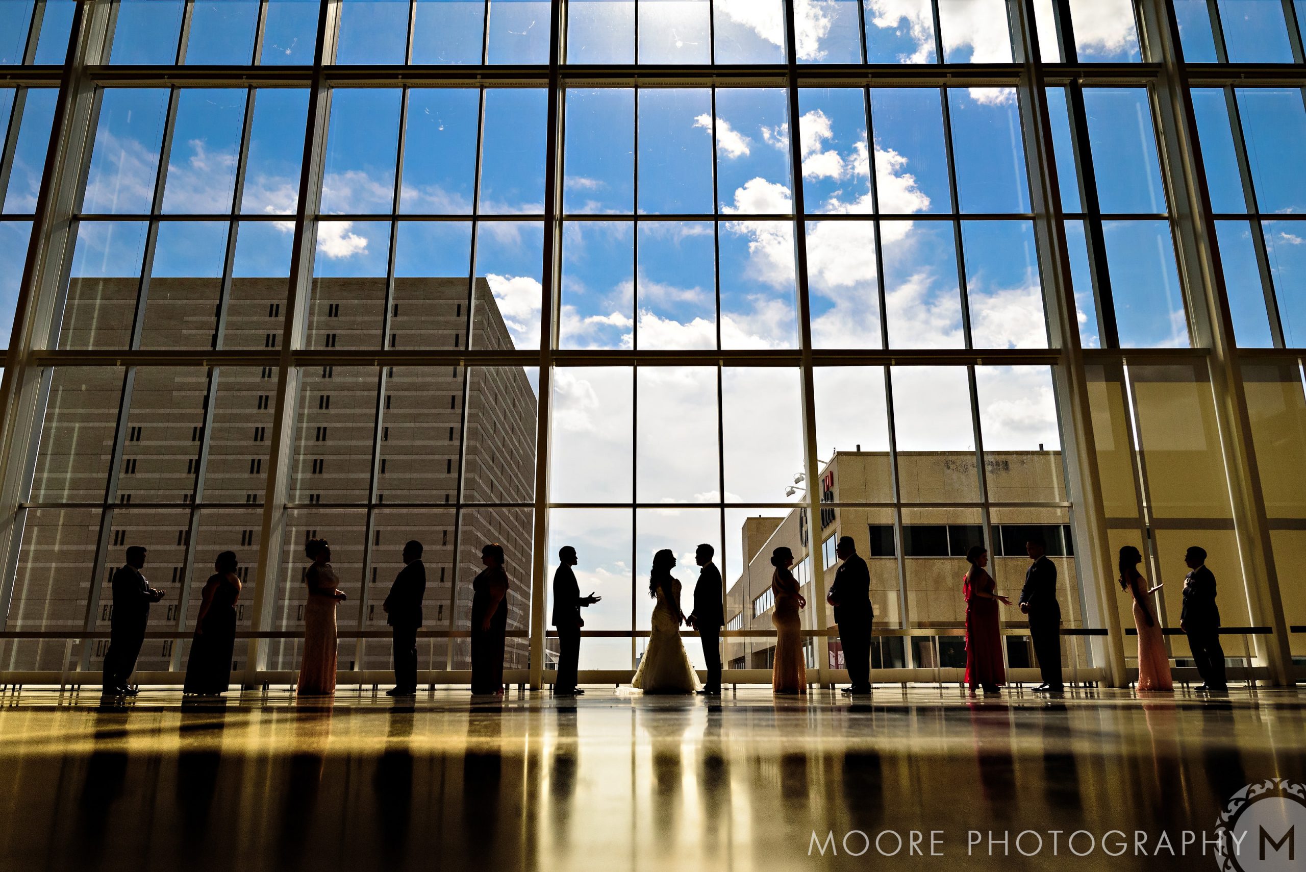 Silhouetted wedding party in Winnipeg venues, large windows frame the bright sky.