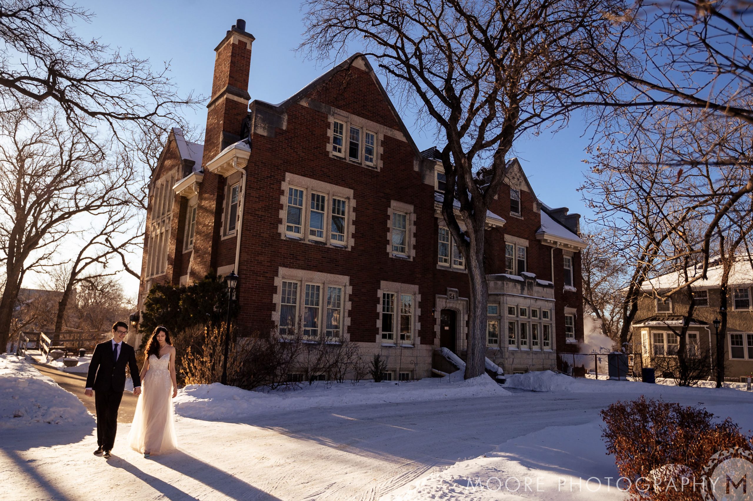 A couple strolls hand-in-hand near a brick house on a sunny, snowy Winnipeg day.