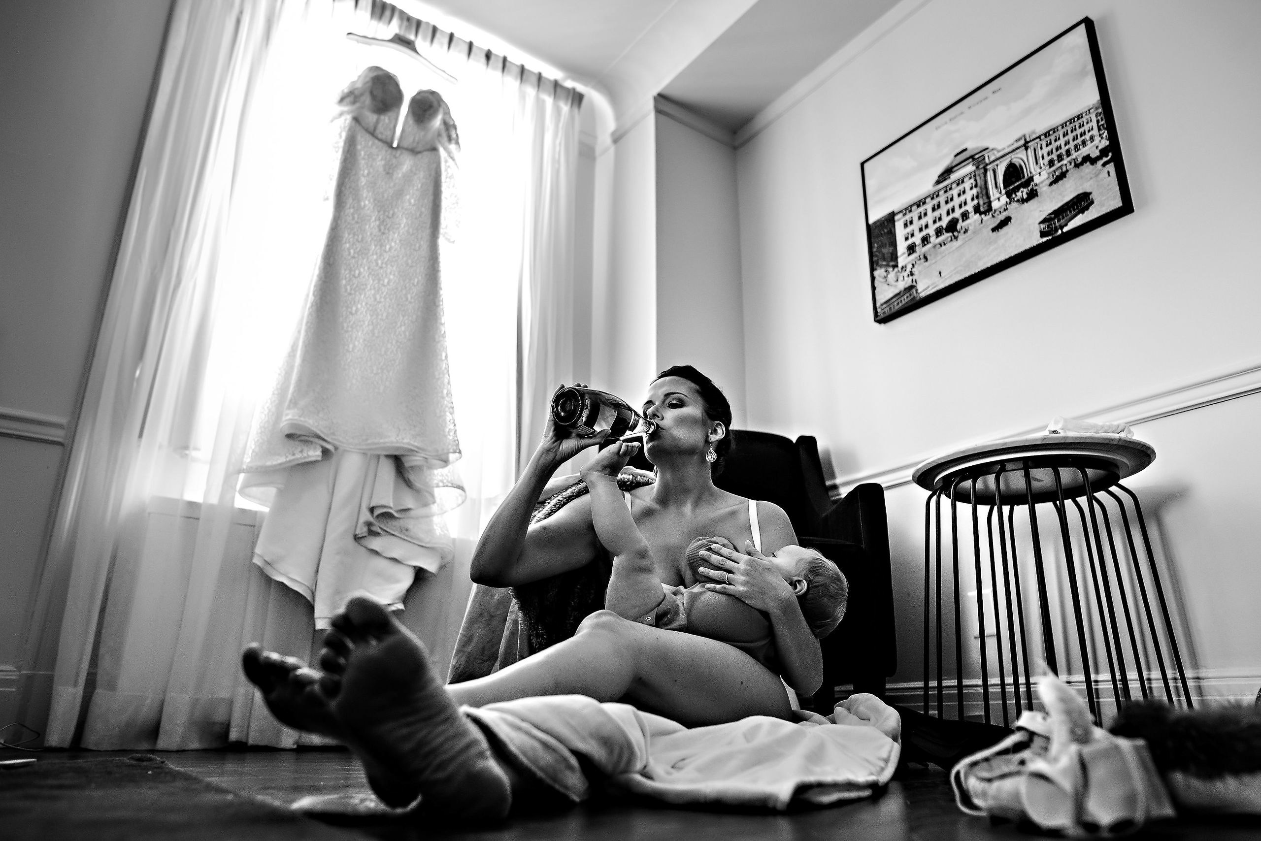 In a cozy Winnipeg wedding venue, a woman breastfeeds, sipping from a bottle, dress nearby.