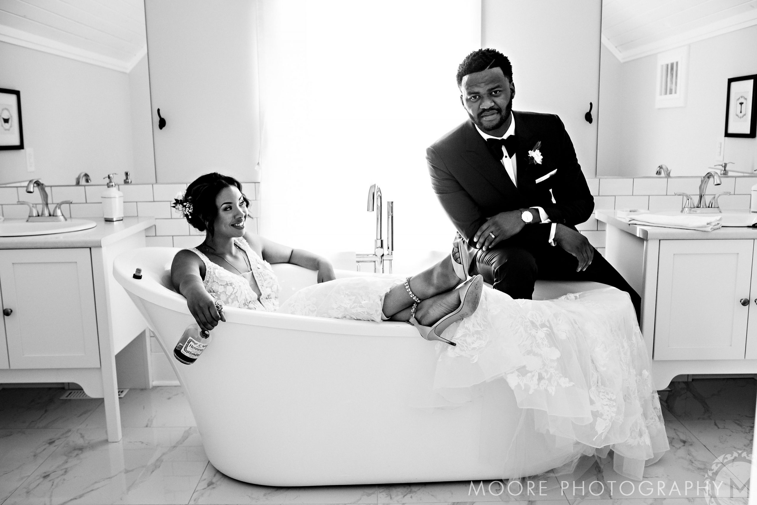 Bride in tub with drink, groom on edge, both smiling; Winnipeg wedding attire.