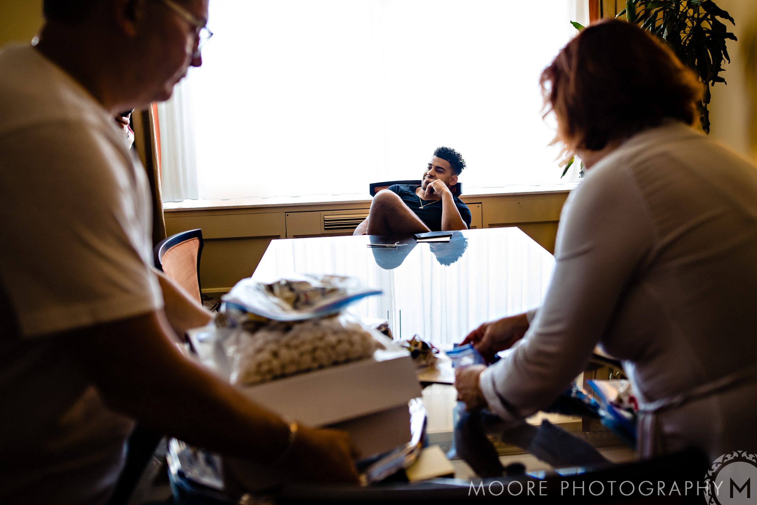 Andrew Harris sits at a table, two stand chatting. Bright room with a large window.