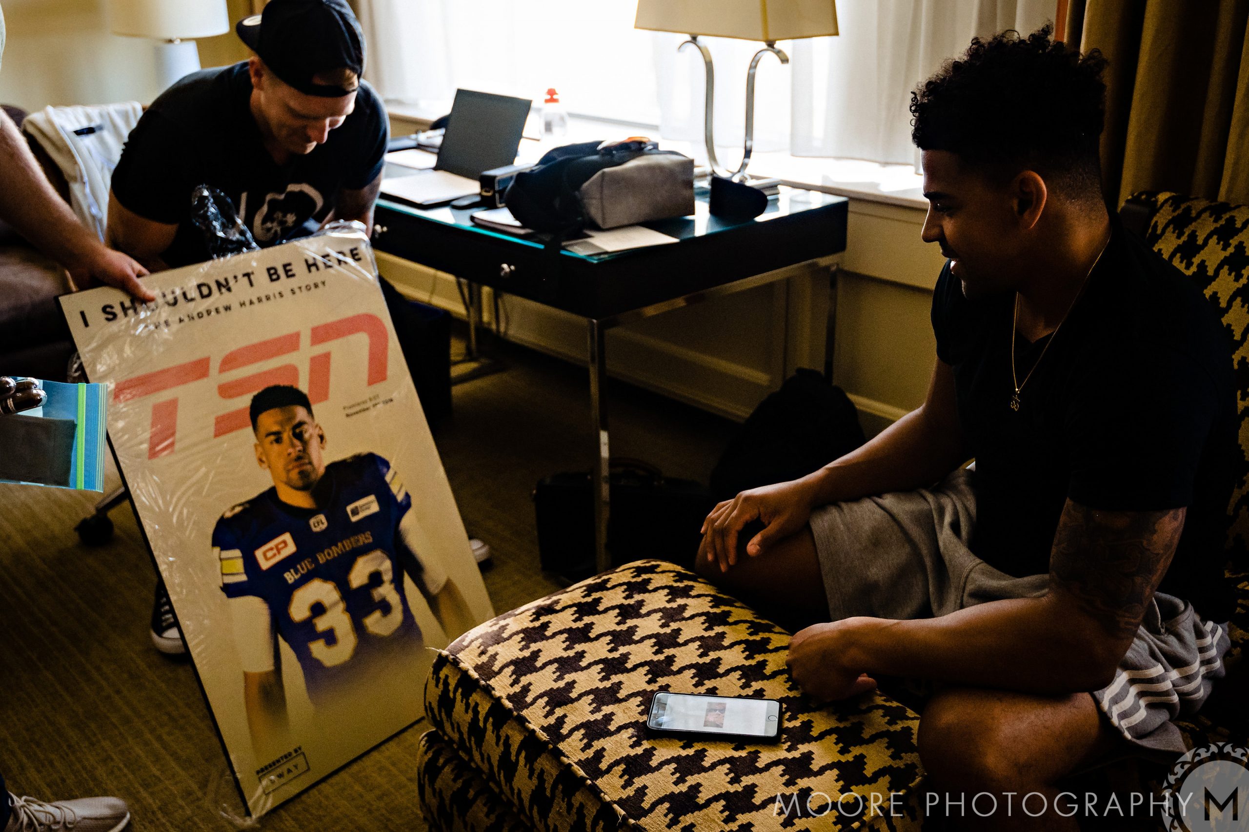 Andrew Harris and a friend sit in a room, one holding a framed sports poster.