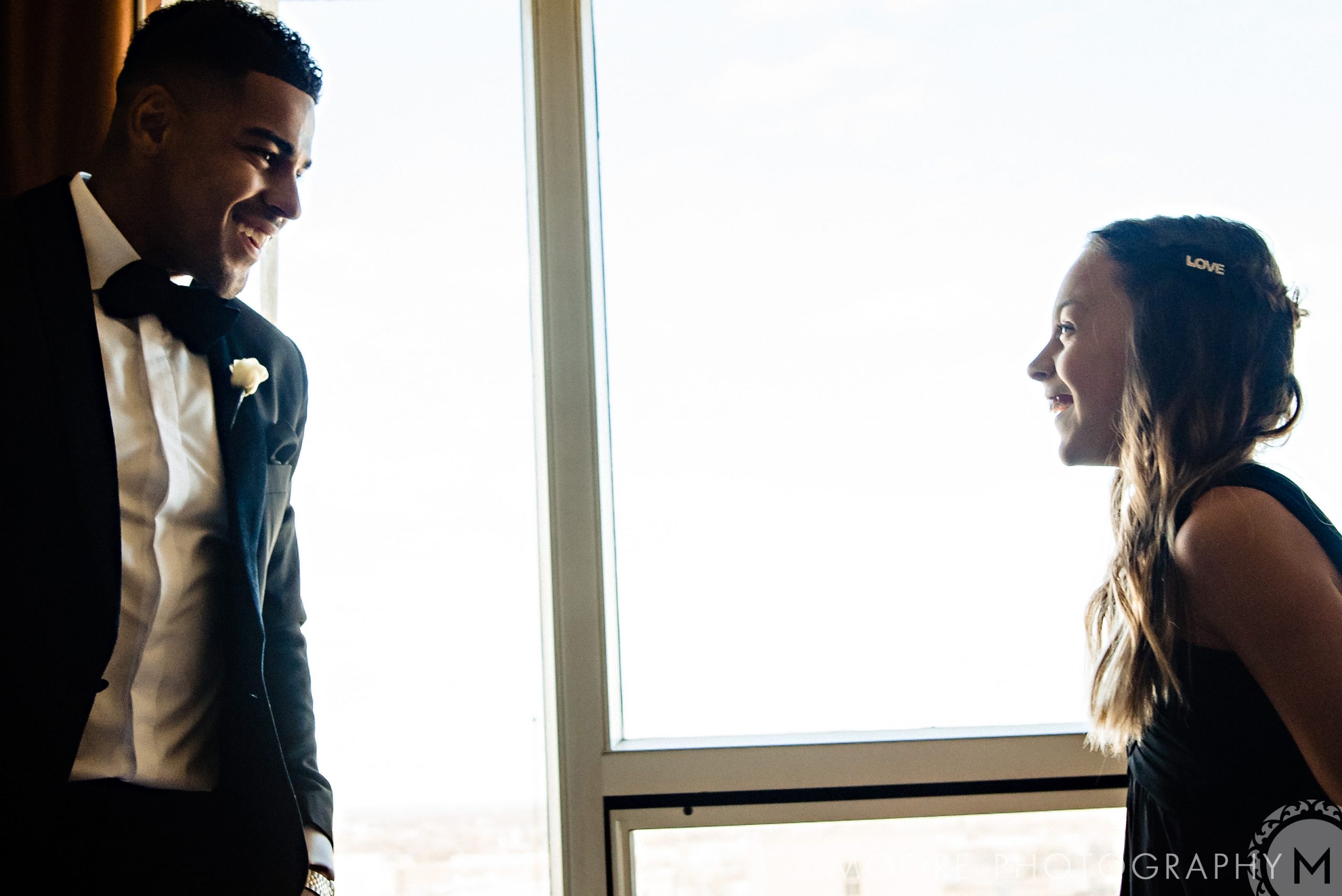 Andrew Harris, in a suit, smiles at a girl by a window with a city wedding view.