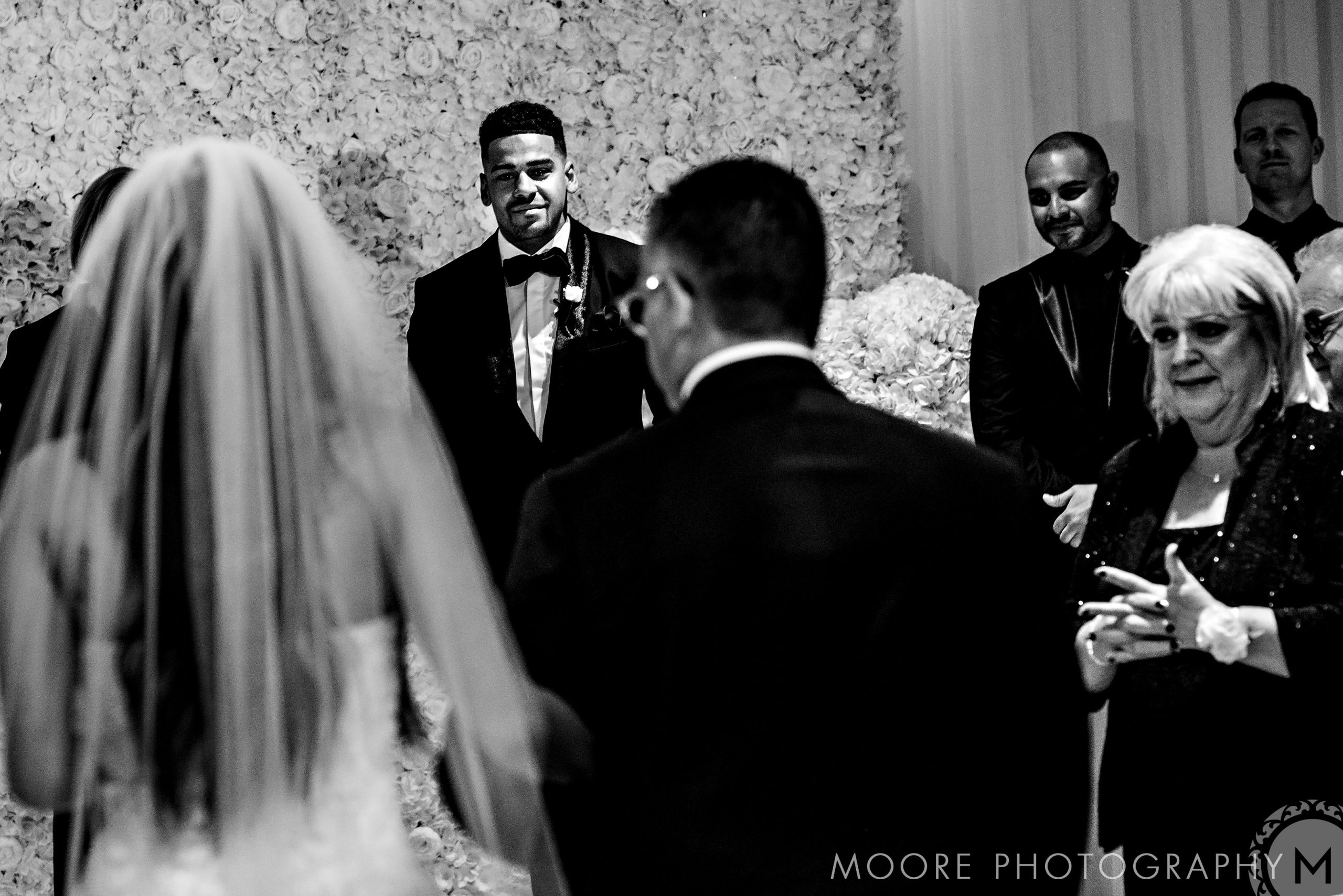 Bride approaches groom, Andrew Harris, in a flower-adorned ceremony; guests watch eagerly.