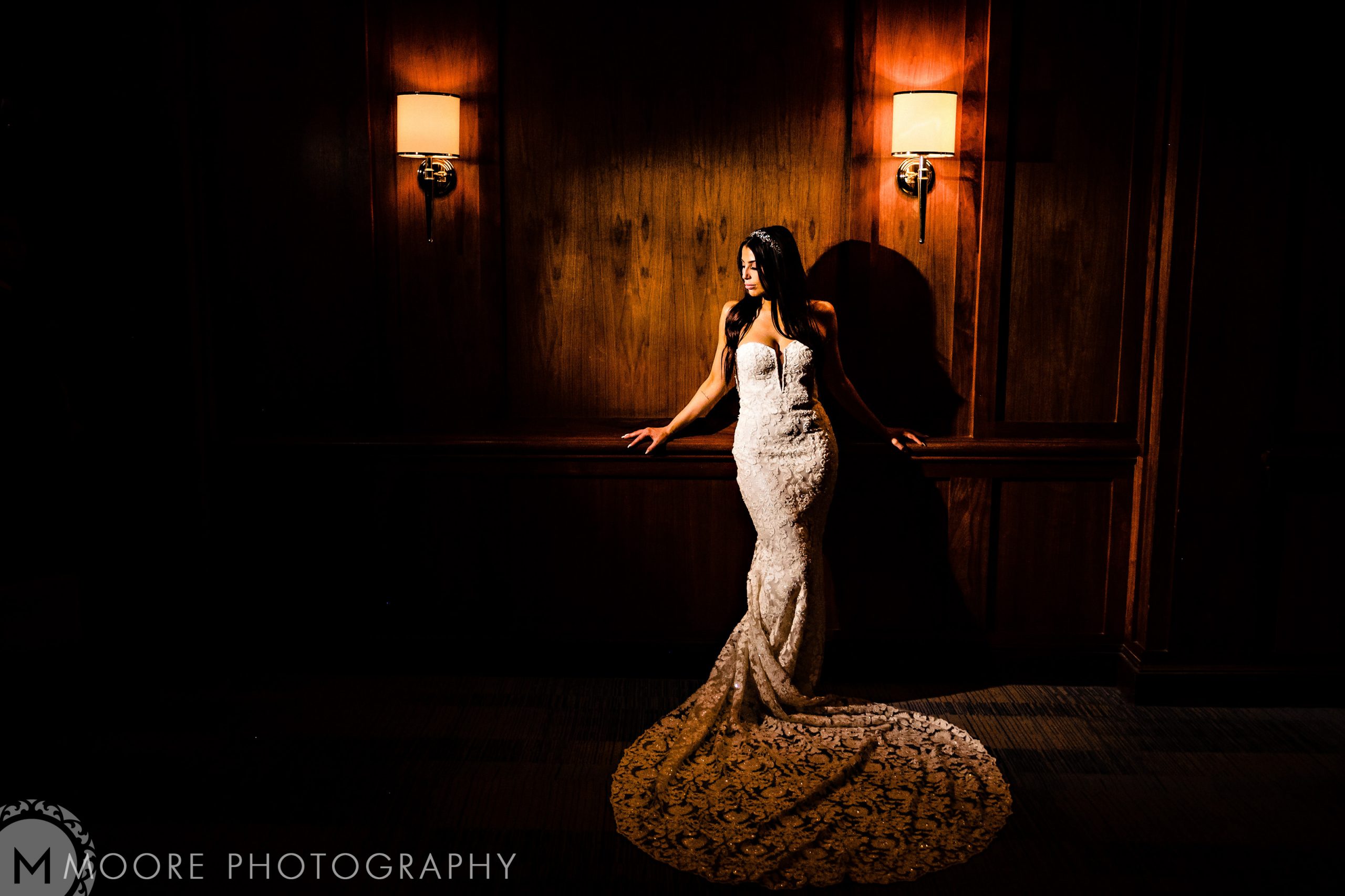 Bride in lace gown stands by dimly lit wooden wall at Andrew and Harris wedding.