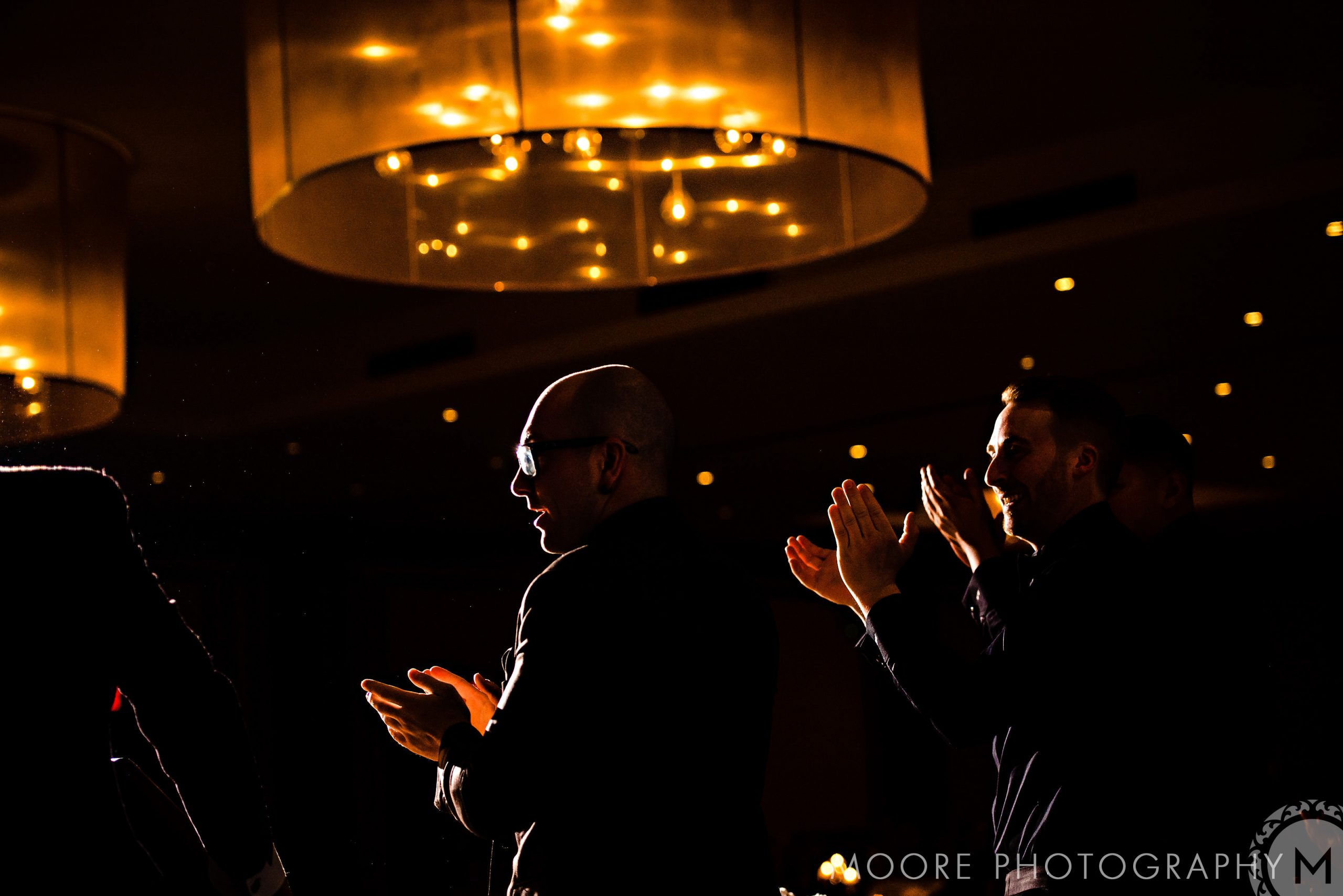 Under warm, glowing chandeliers, guests clap at Andrew Harris's wedding.