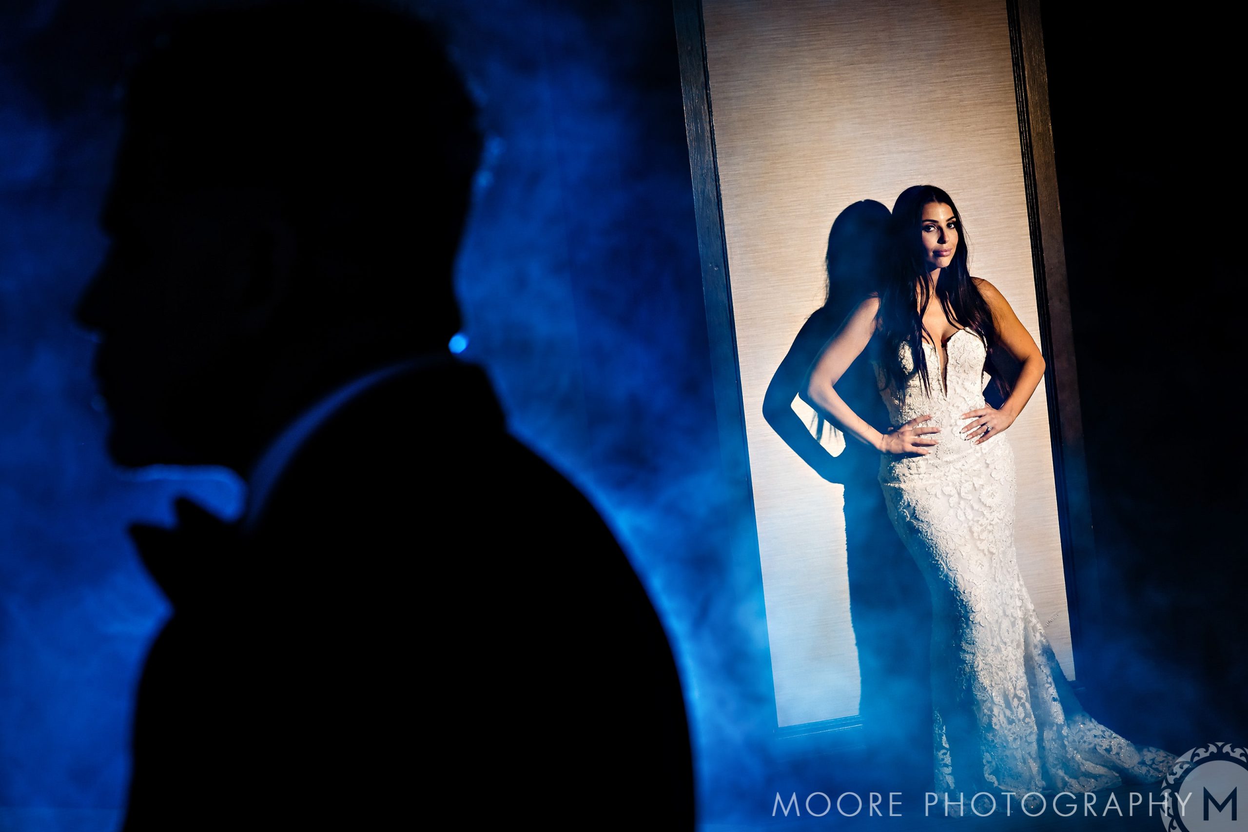 Bride posing in lighted area, shadowy groom foreground, blue mist at an Andrew Harris wedding.