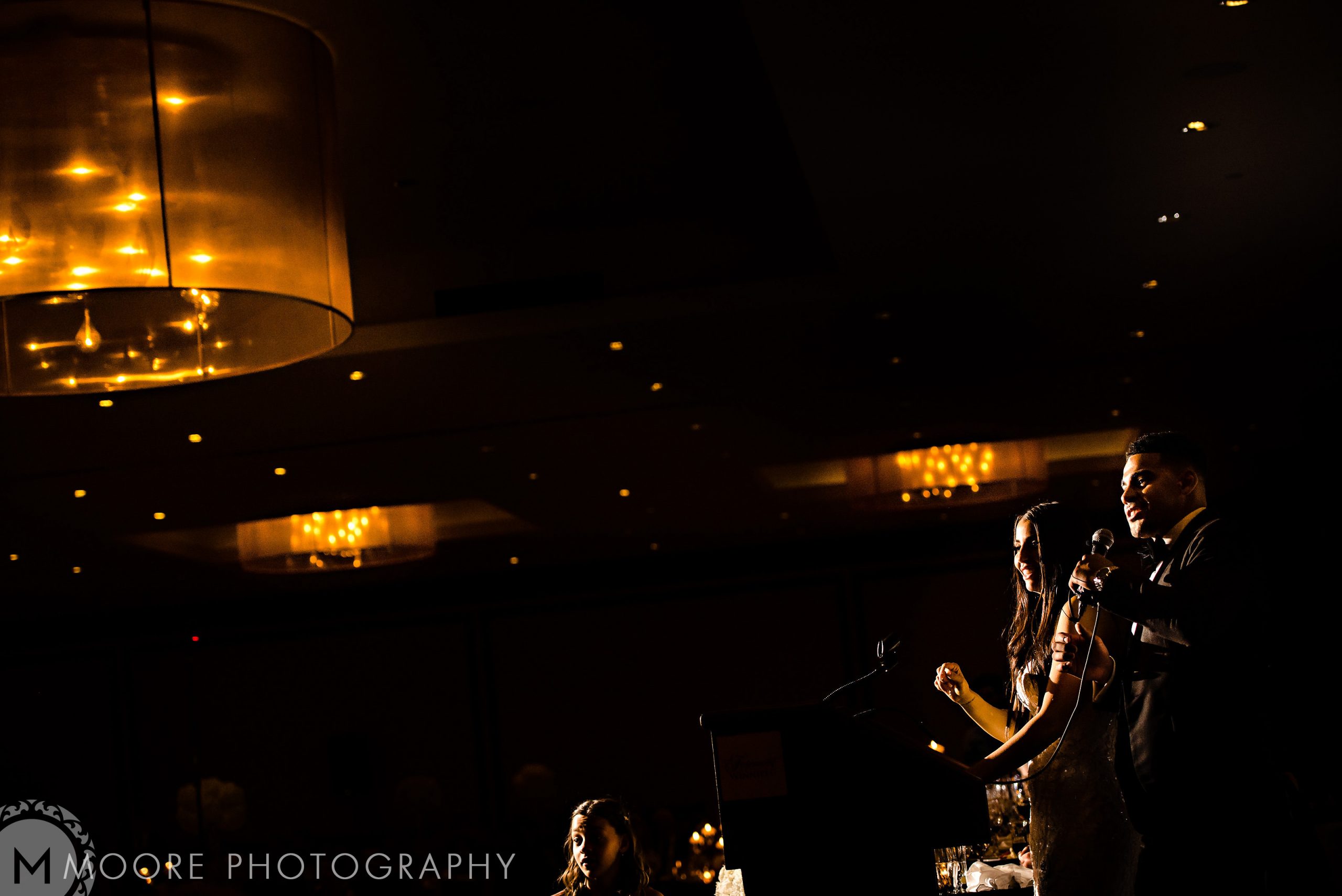 A speaker at a dimly lit Andrew Harris wedding with a large ceiling light above.