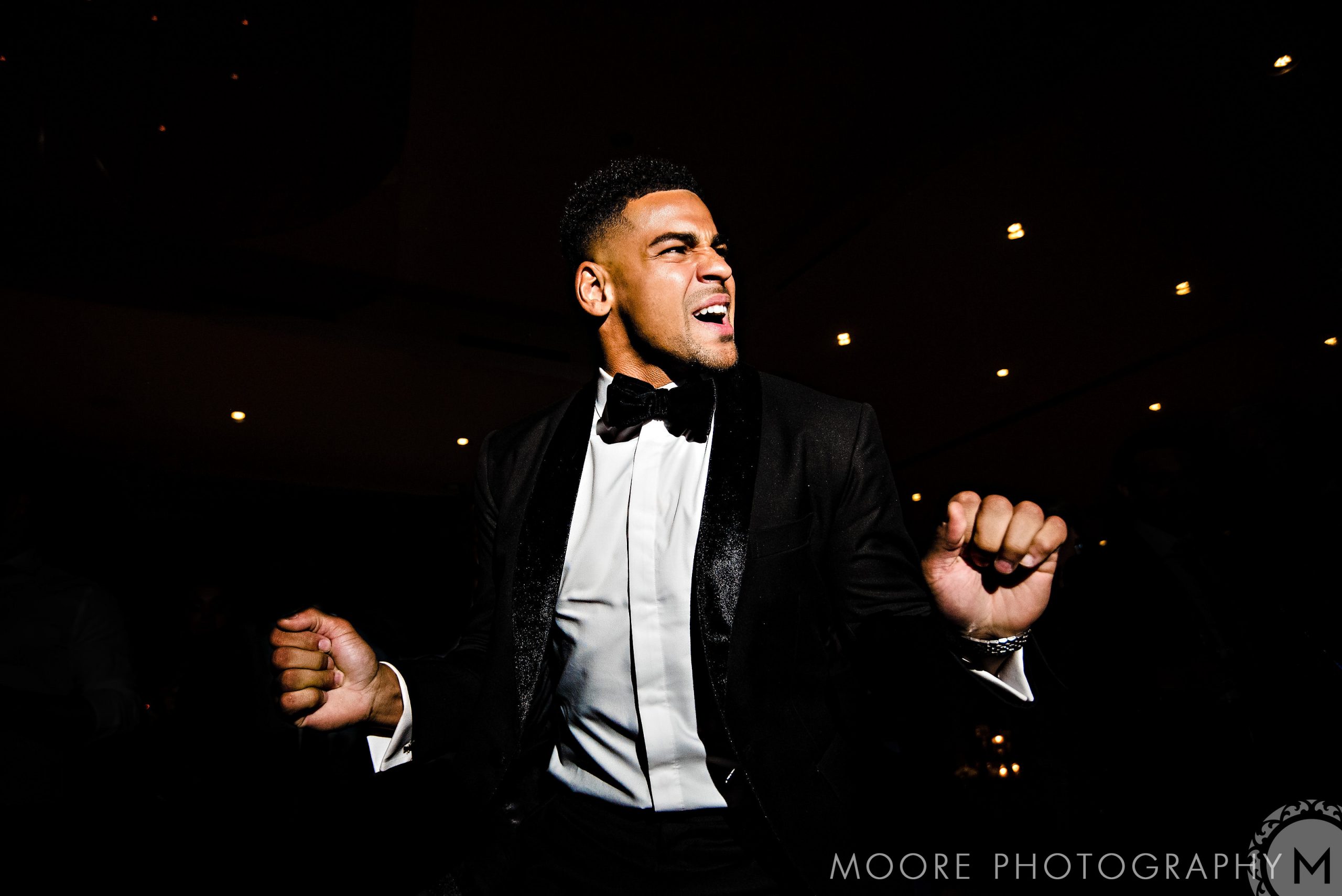 Andrew Harris dancing energetically in a tuxedo under bright wedding lights.