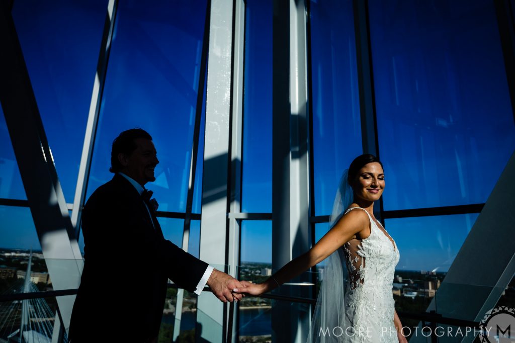 Bride and groom holding hands inside a sunlit Winnipeg wedding venue, sky blue outside.