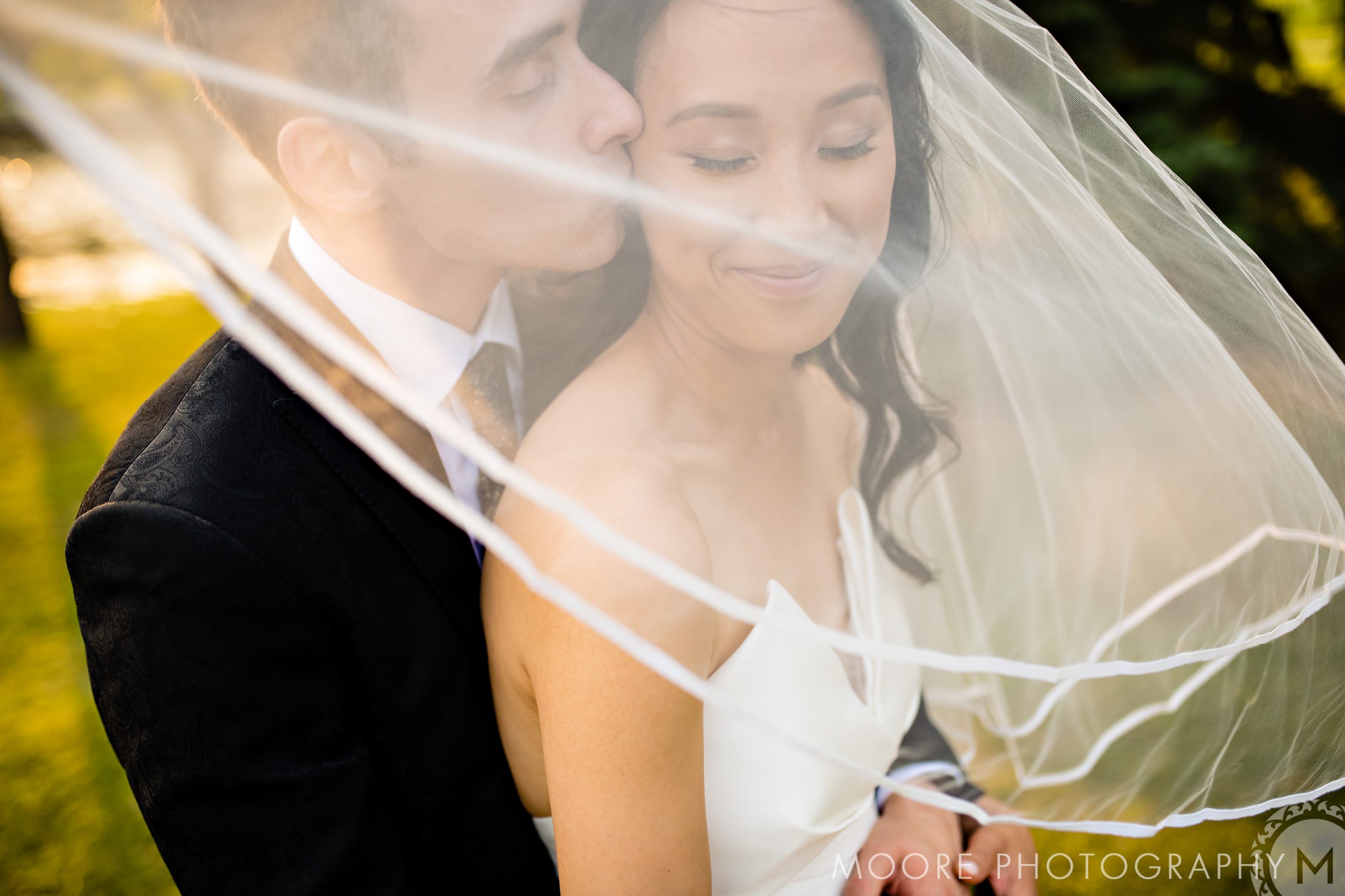 A couple embraces under a veil in a sunlit Winnipeg wedding garden.