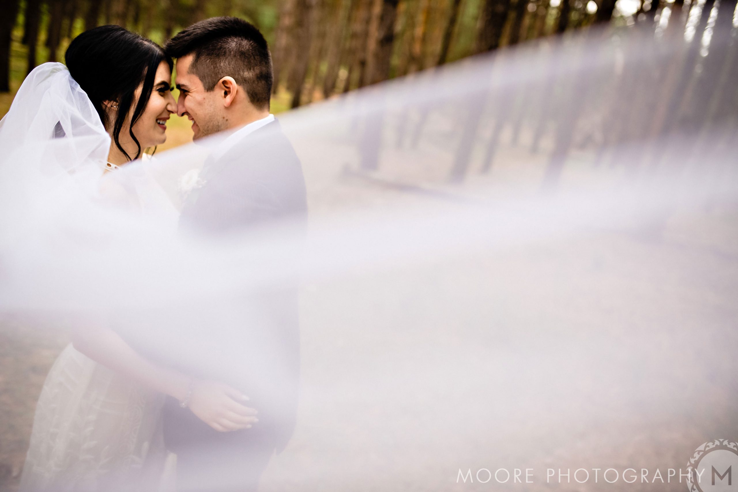Bride and groom smile in a forest, white veil flowing at a Winnipeg wedding venue.