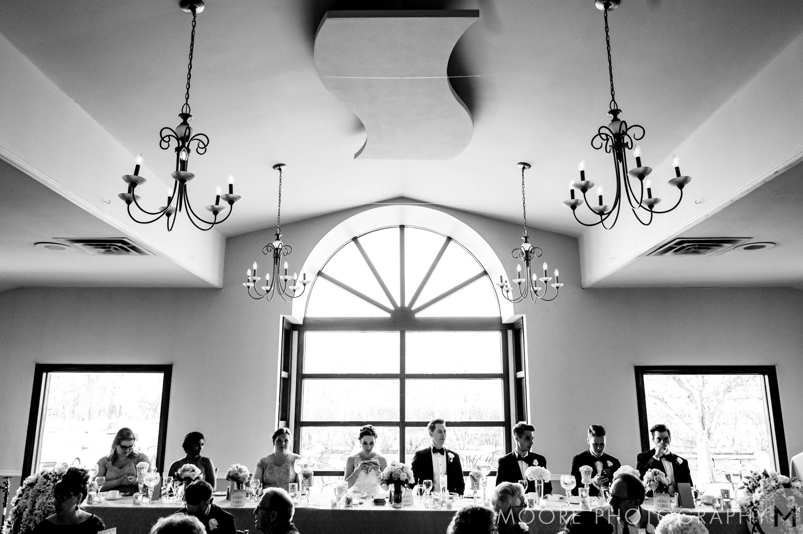 The wedding party gathers at a long table in a Winnipeg venue with arched windows and chandeliers.