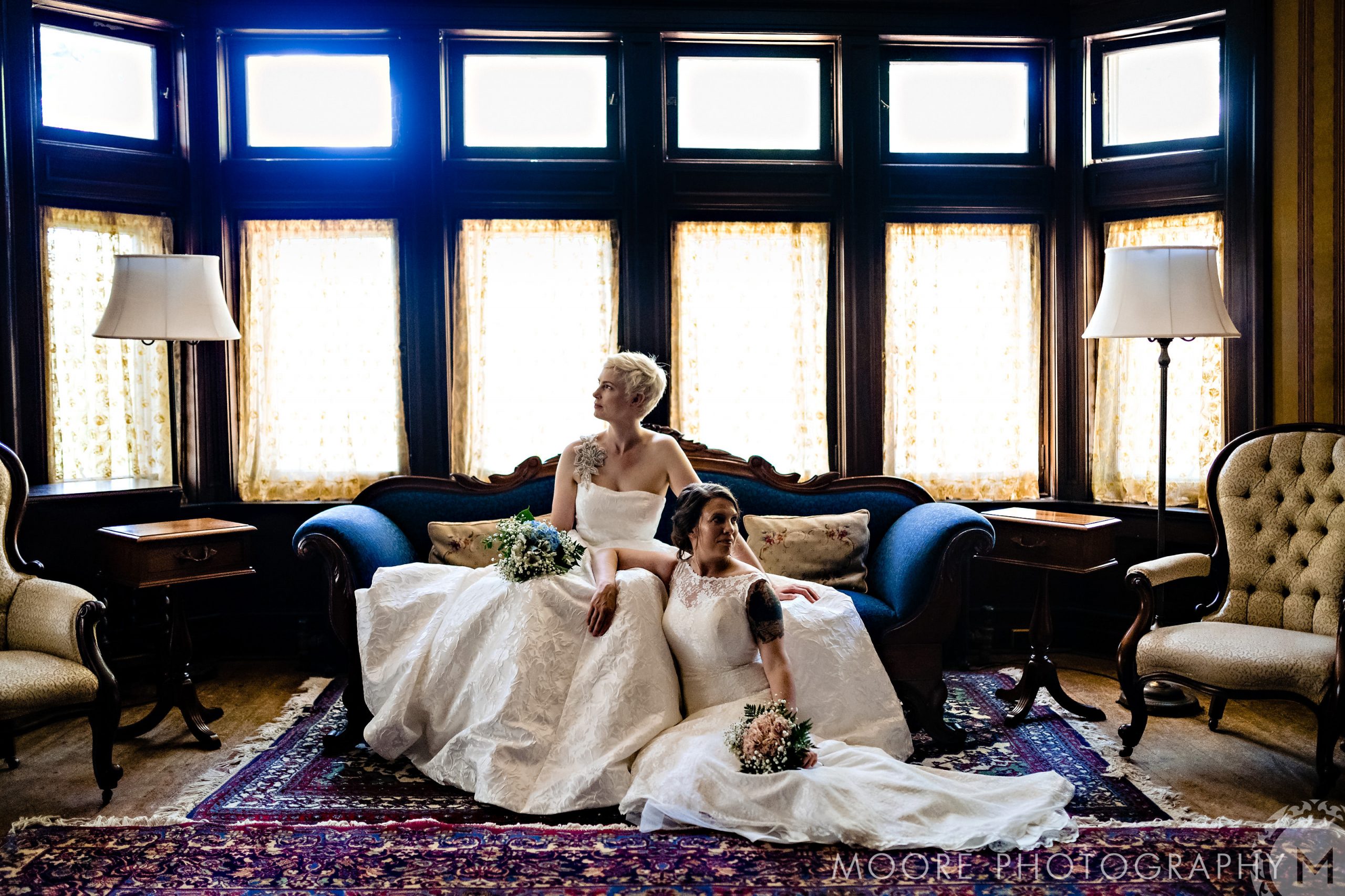 Two brides in wedding dresses pose on a vintage sofa at a charming Winnipeg wedding venue.