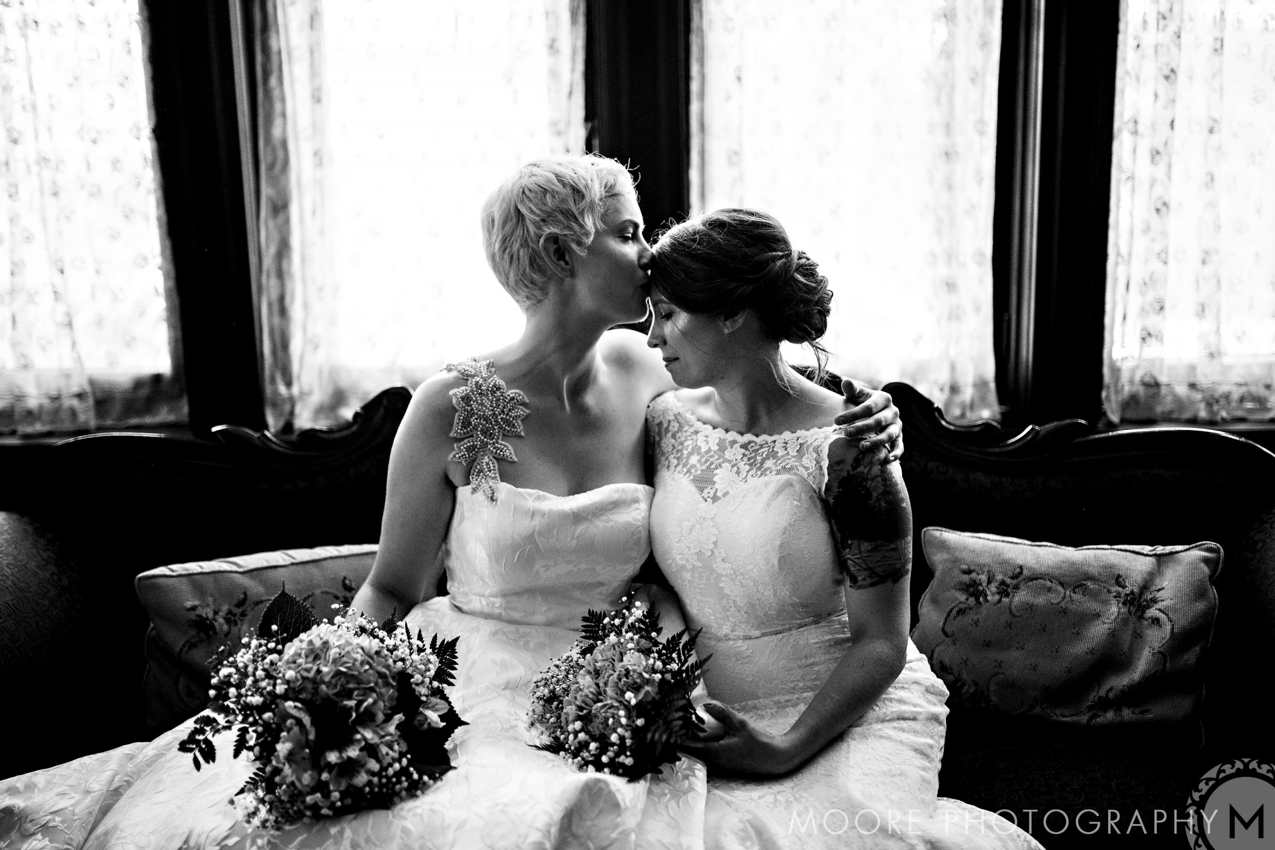 Two brides on a sofa, one kissing the other's forehead, holding bouquets. Winnipeg wedding bliss.