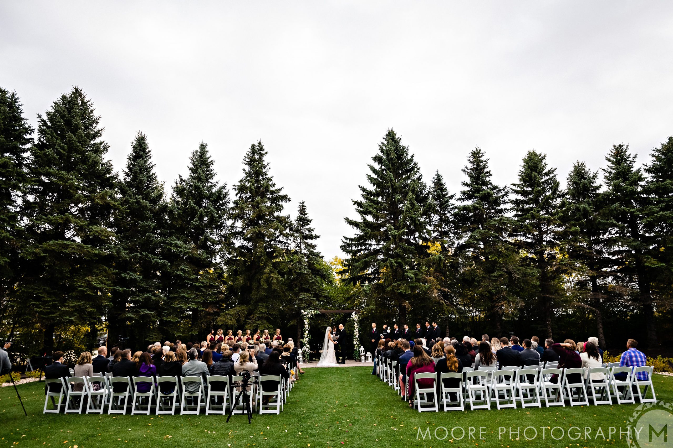 Winnipeg wedding venues host outdoor ceremonies on lawns, surrounded by tall evergreen trees.