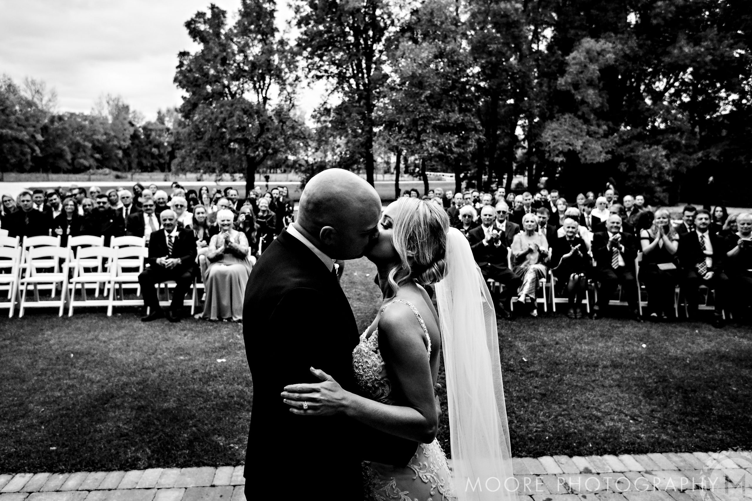 Bride and groom kiss outdoors at Winnipeg wedding, guests seated nearby.