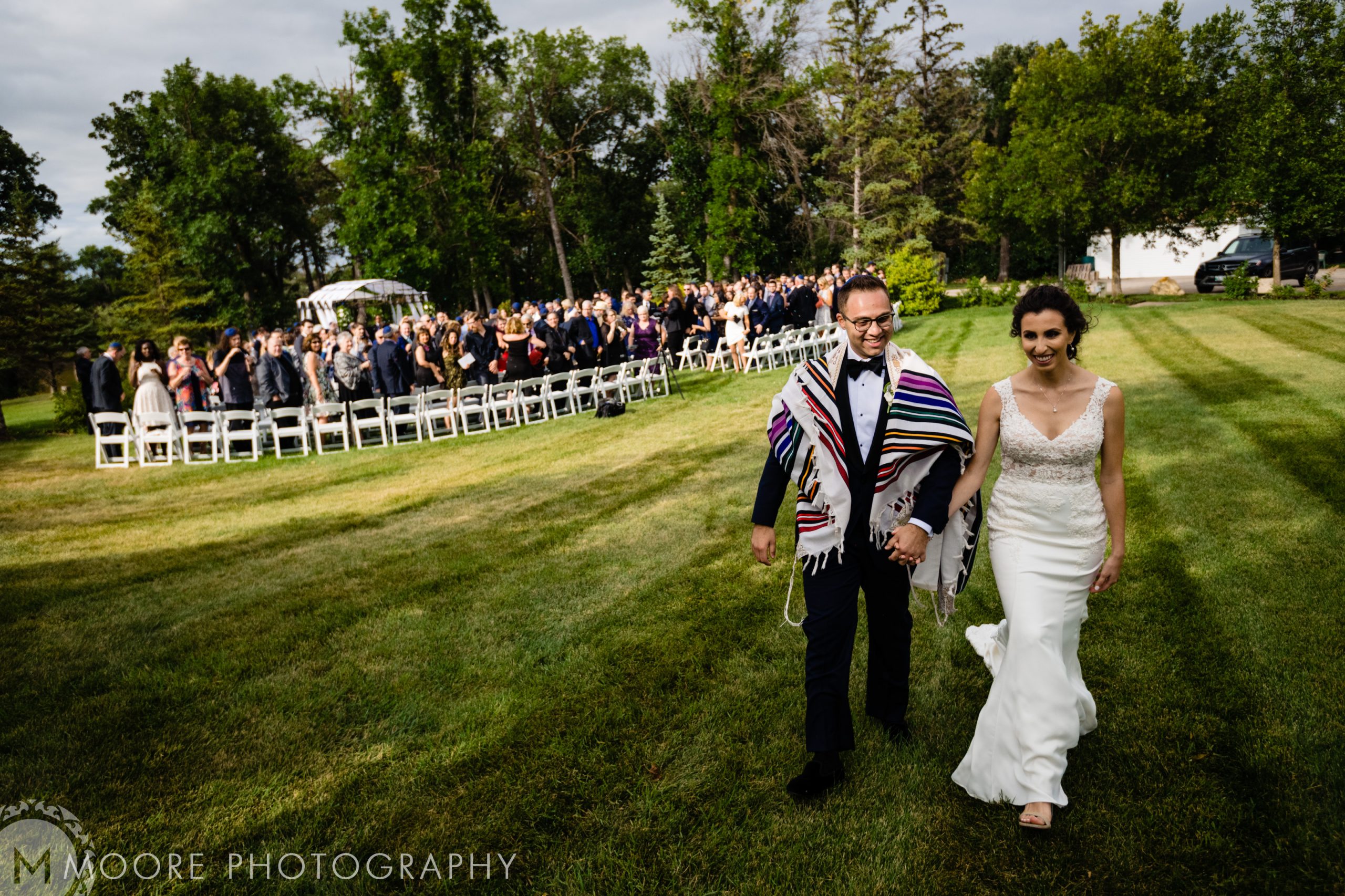 A couple strolls hand-in-hand at a lively Jewish wedding ceremony.