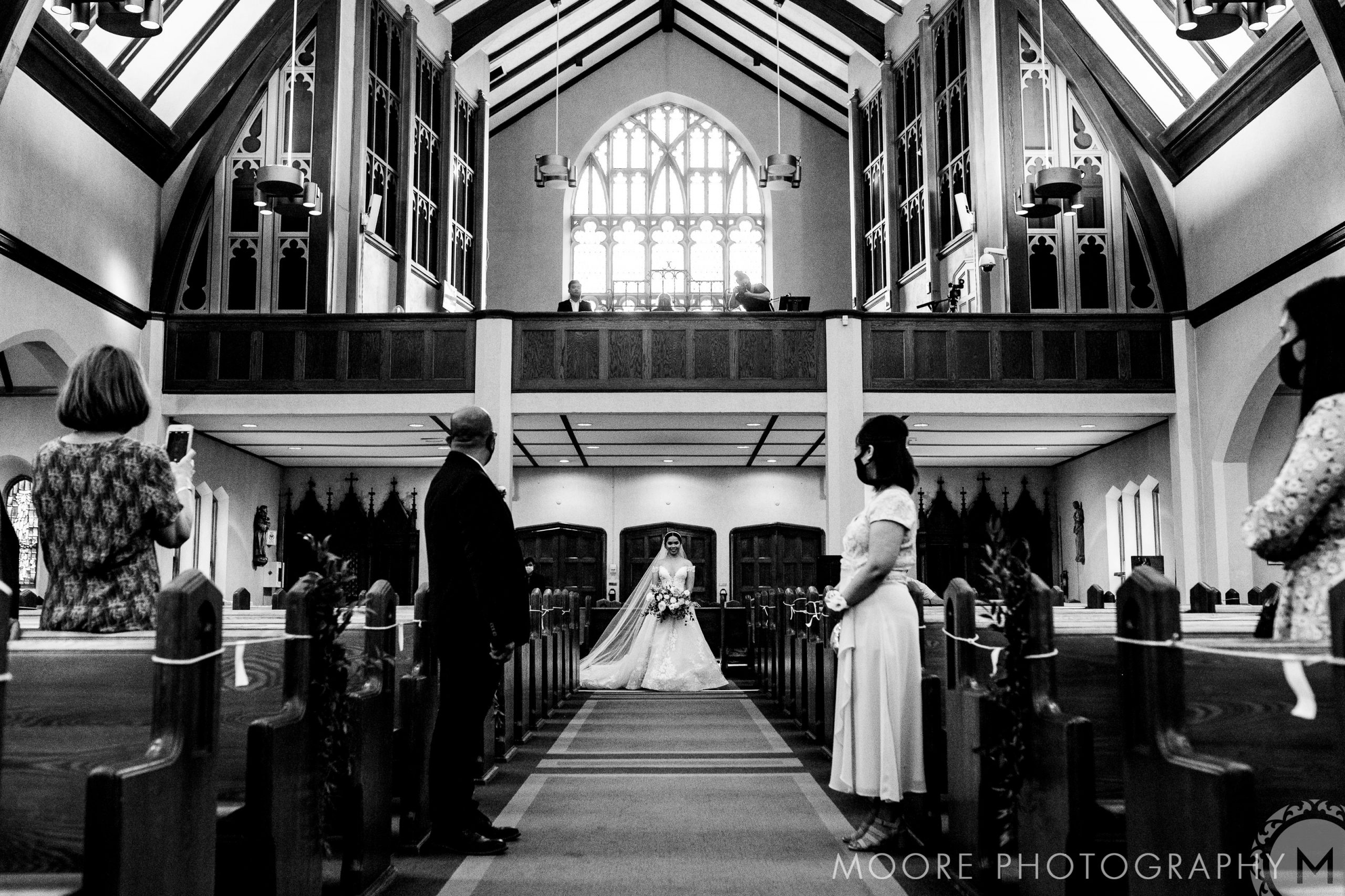 A bride walks down the aisle of a large, ornate church with high ceilings and stained glass windows, accompanied by people standing on both sides of the aisle, watching her. The image is in black and white, and several people are taking photos. "Moore Photography" is seen in the bottom right corner.