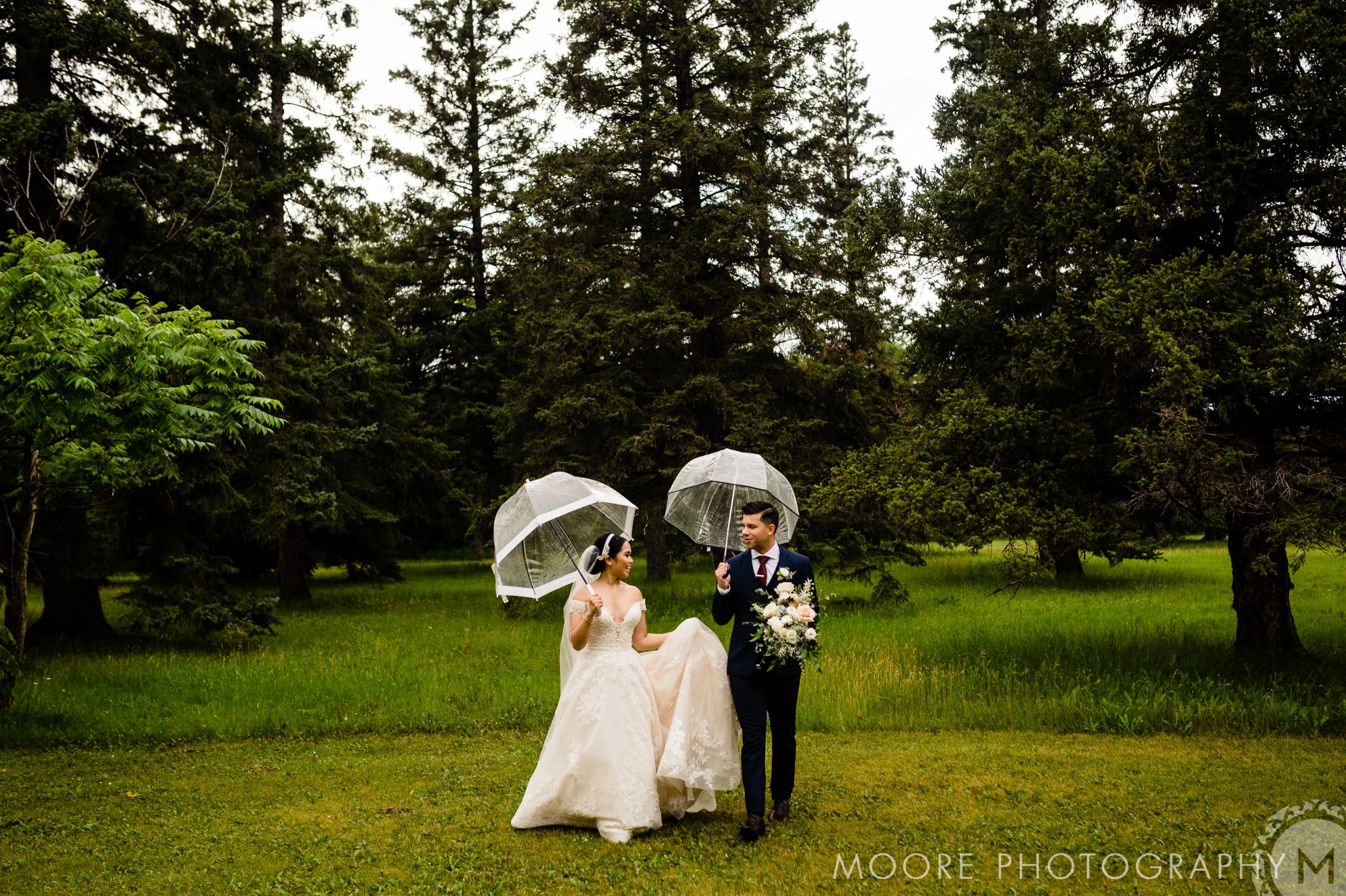 A bride and groom walk hand in hand on a grassy path surrounded by tall trees. Both are holding transparent umbrellas. The bride wears a white gown, and the groom dons a dark suit while carrying a bouquet. The image is credited to Moore Photography.