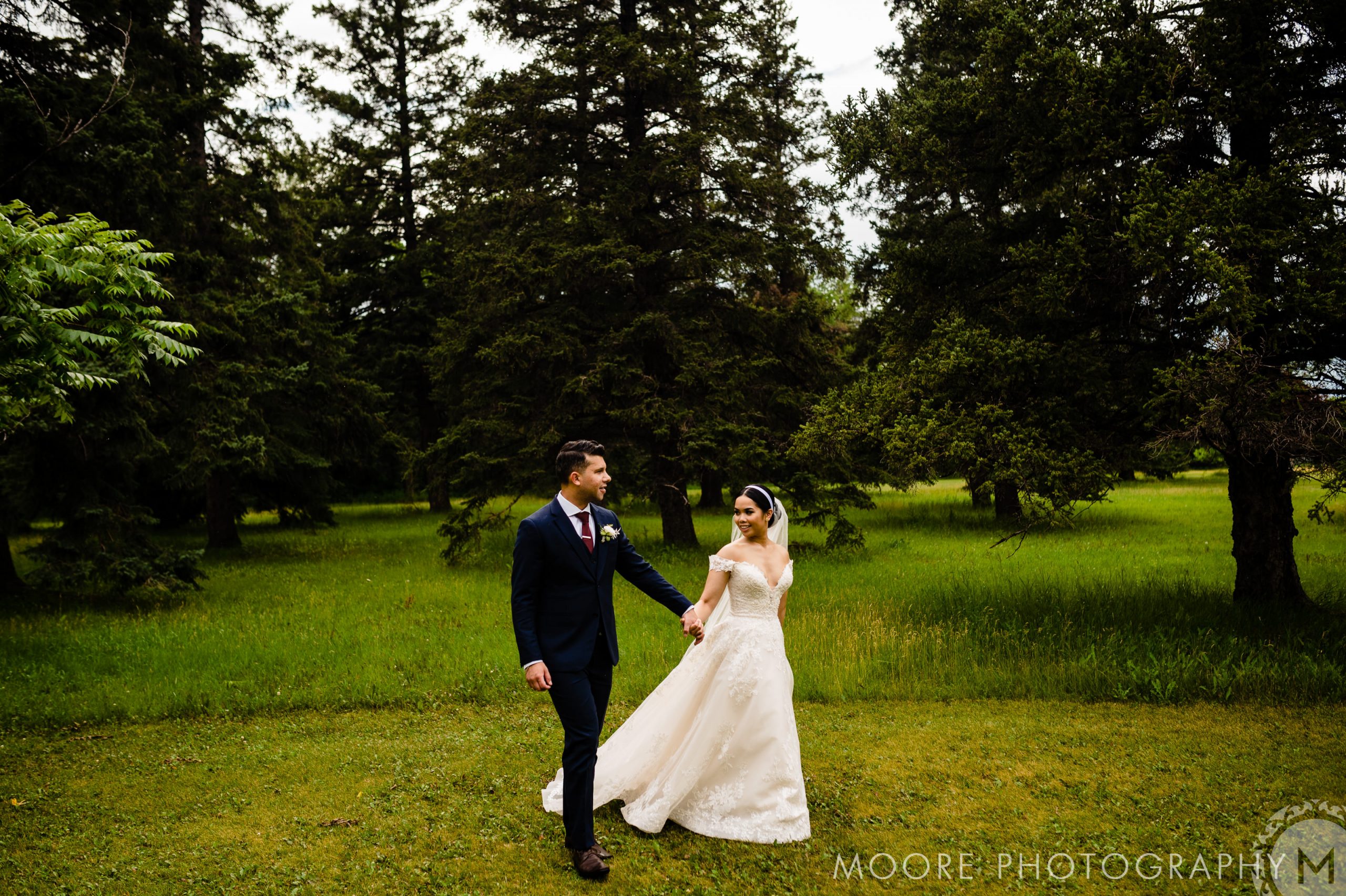 A bride in a white wedding dress and groom in a dark suit hold hands and walk through a grassy area surrounded by trees. The bride smiles while looking at the groom. The scene is set outdoors, with lush greenery in the background. Text at the bottom reads "MOORE PHOTOGRAPHY.