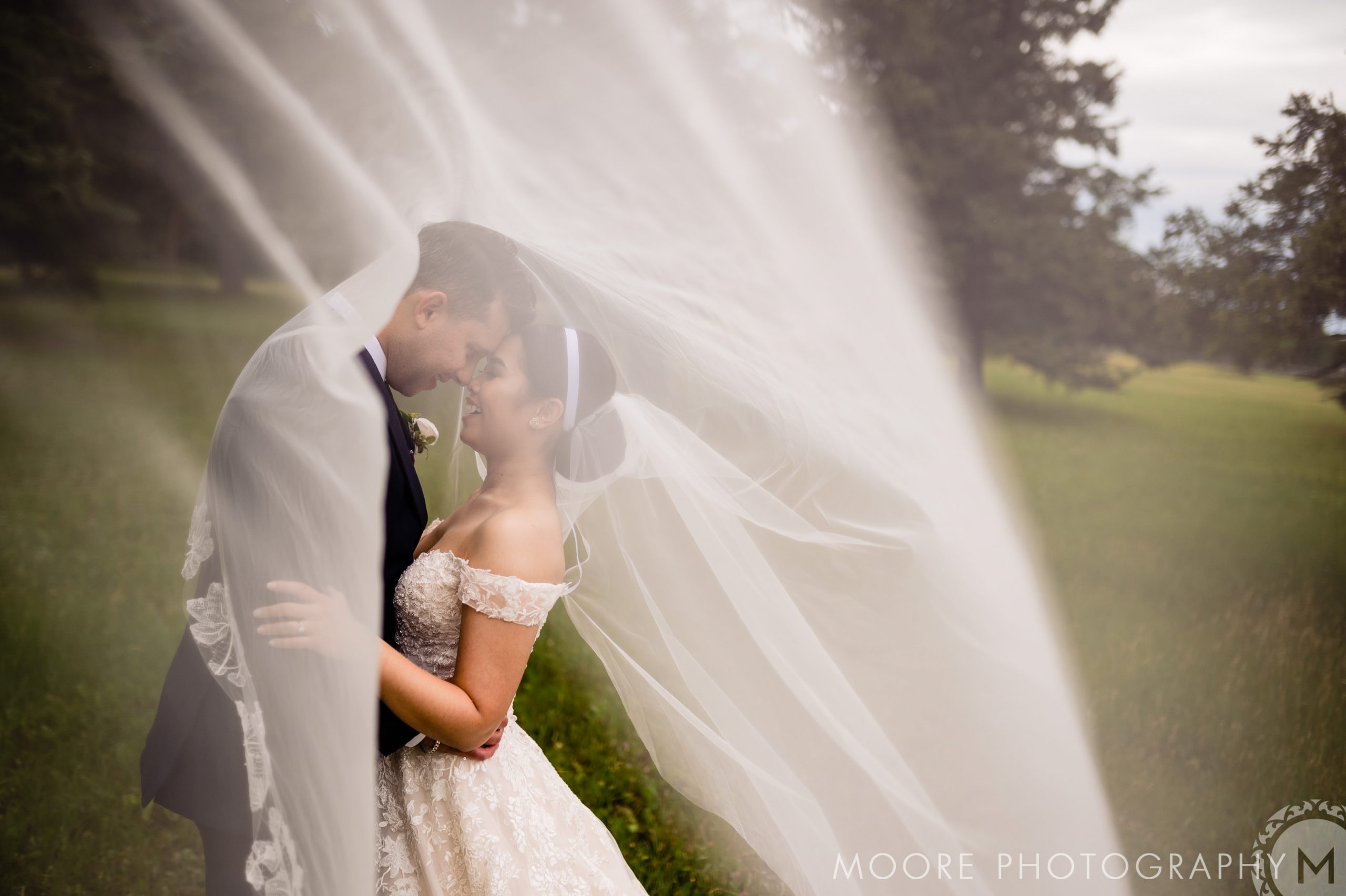 A bride and groom embrace outdoors, under a flowing white veil that envelopes them. The bride is in a white lace dress and the groom in a dark suit. They stand on green grass with trees in the background. The photo is by Moore Photography, as noted in the corner.