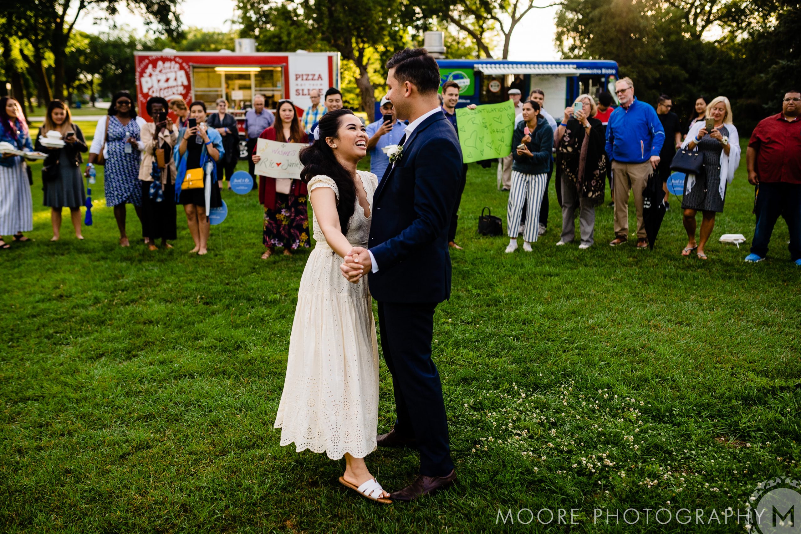A couple in wedding attire dances on a grassy lawn, surrounded by a group of people cheering and holding signs. In the background, there are food trucks and trees. The photo is by Moore Photography.