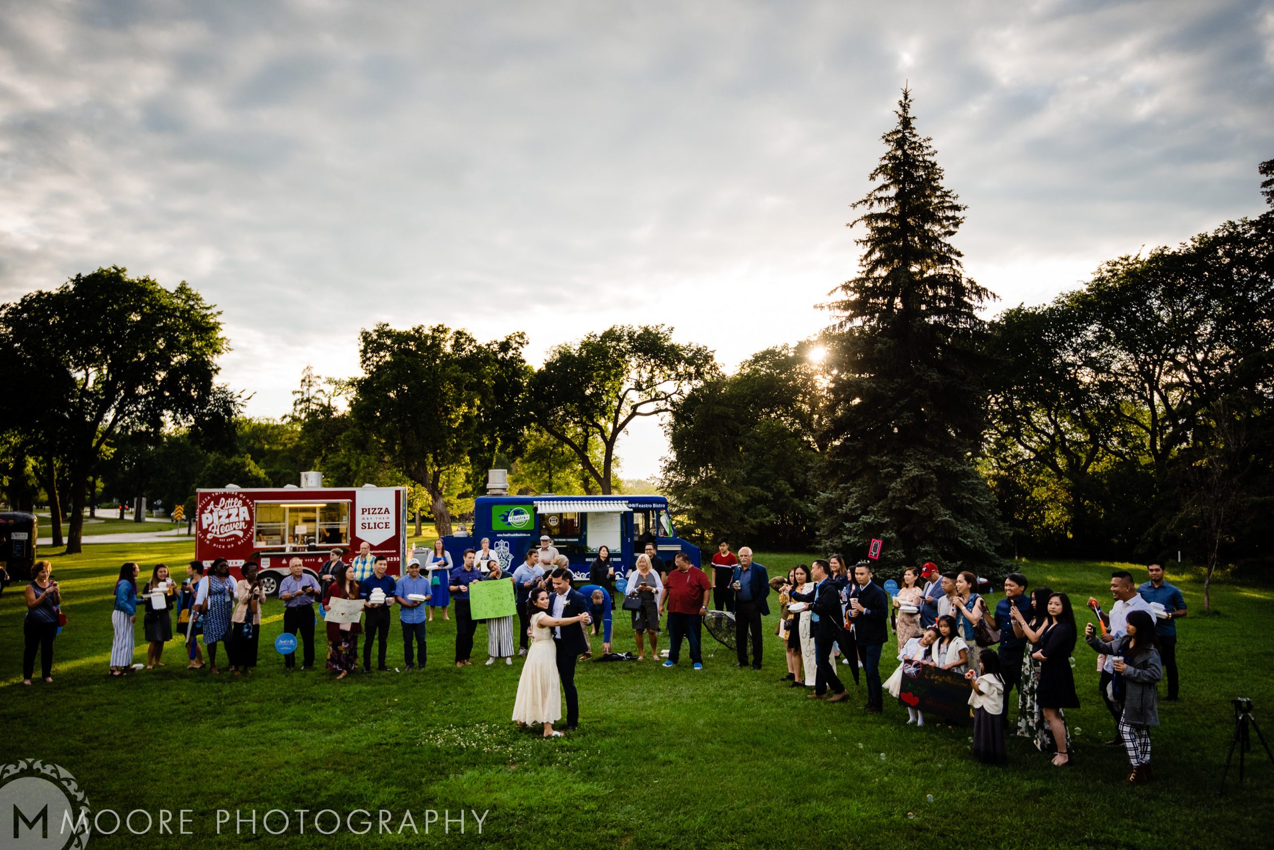 A wedding reception at a Winnipeg park with food trucks and guests on a grassy area.