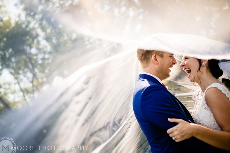 Bride and groom laugh under veil at sunlit Winnipeg wedding venue.