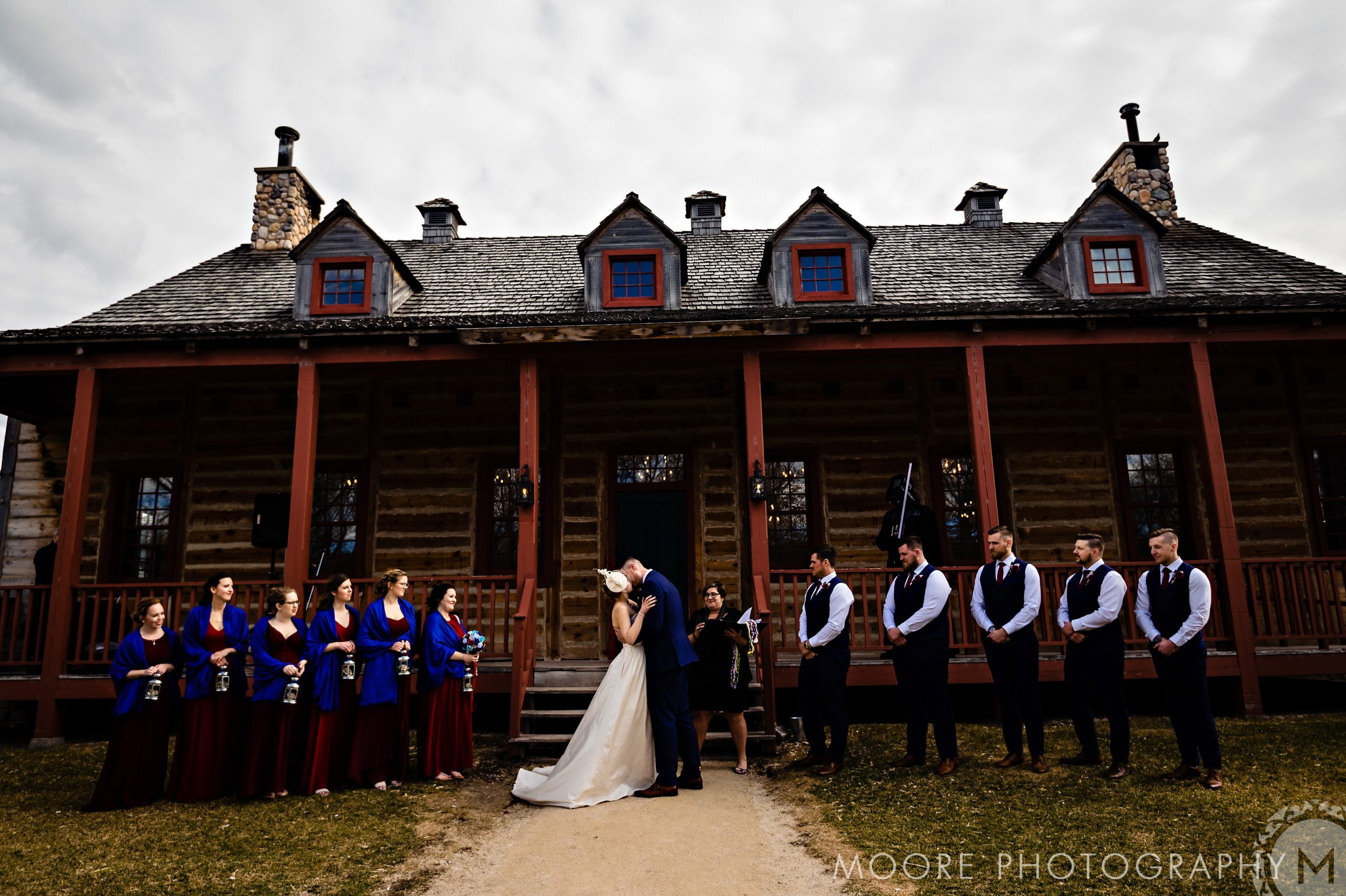 Bride and groom kiss at a rustic Winnipeg wedding venue, wedding party standing by.
