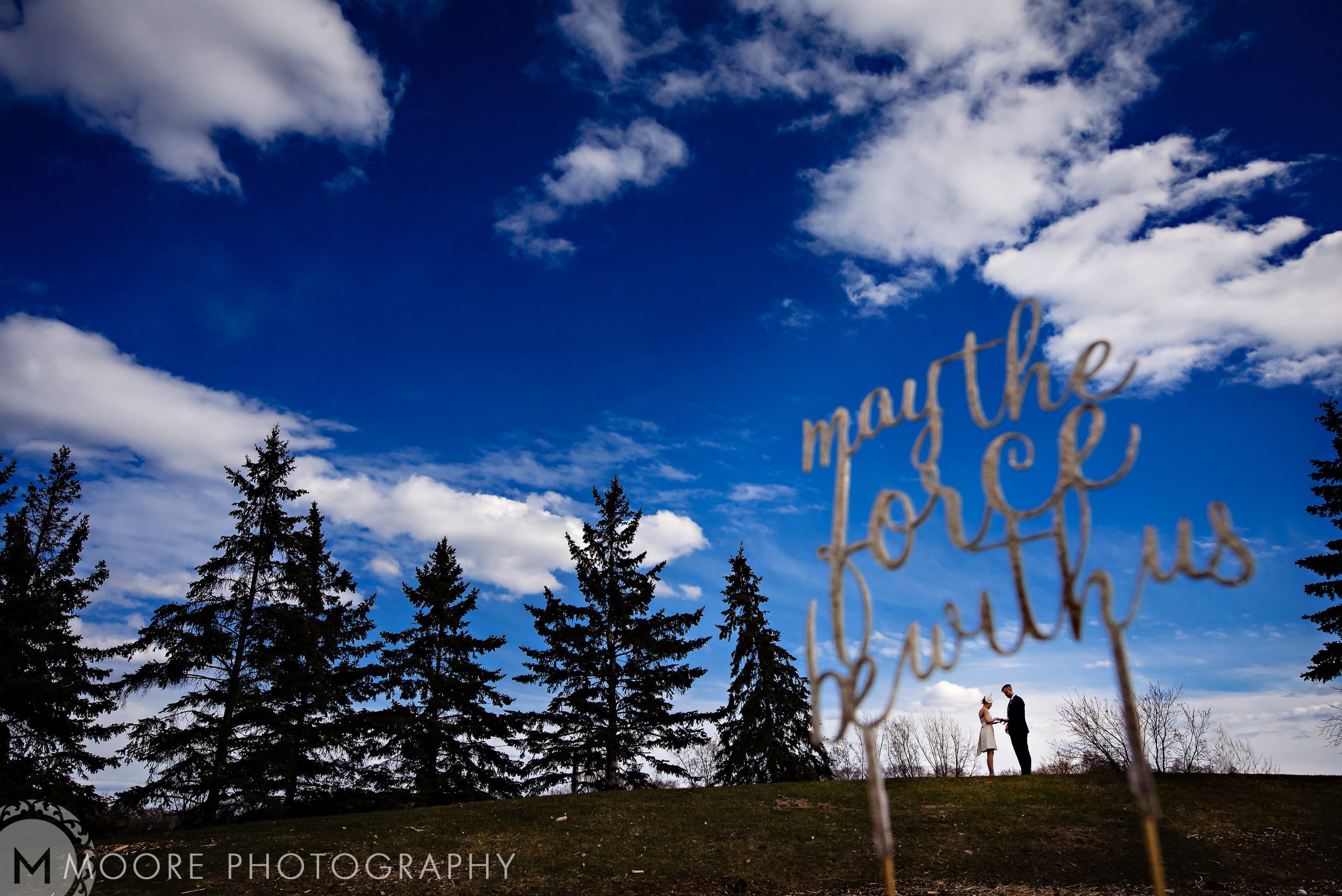 Silhouetted couple on hill, "May the force be with us," blue sky, clouds, pine trees at Winnipeg venue.