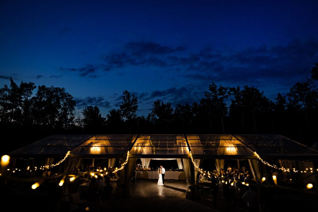 A nighttime outdoor wedding reception with a tented area decorated with string lights, captured brilliantly by our wedding photographer. A couple dances alone on the dance floor, while guests watch from their tables inside the illuminated tent. The sky above is dark blue, with trees silhouetted in the background.