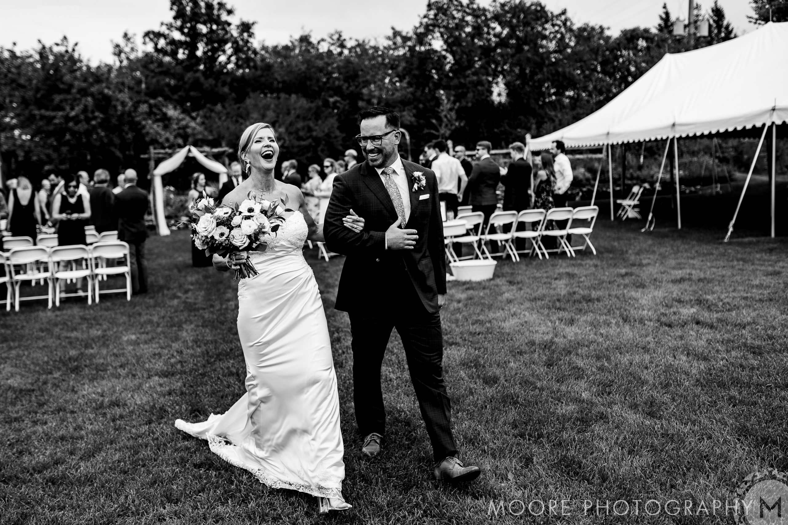 Joyful bride and groom walking down aisle outdoors at Winnipeg wedding venue, in black and white.