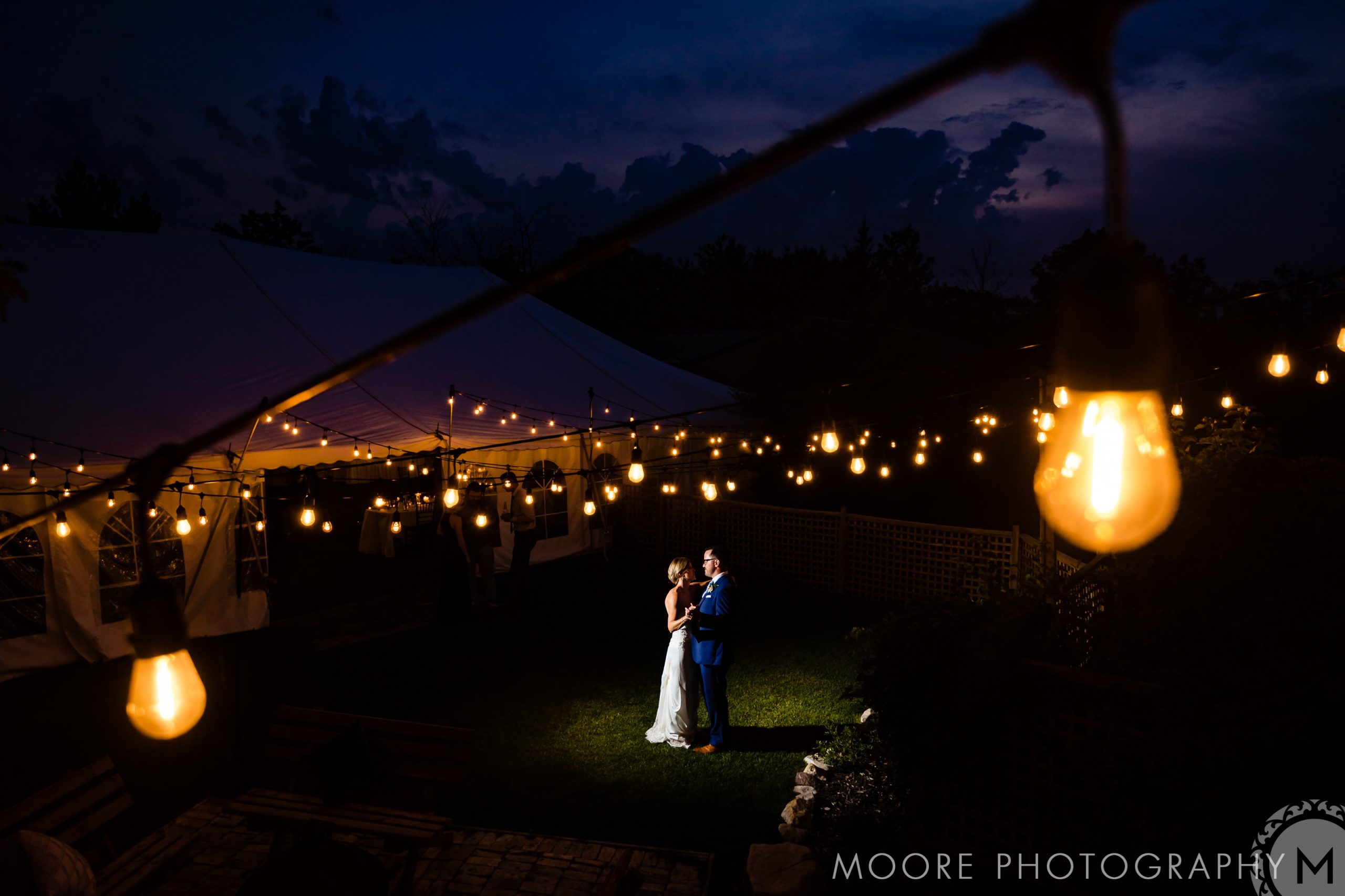 Couple dances under string lights at a Winnipeg wedding venue, dark sky in the background.