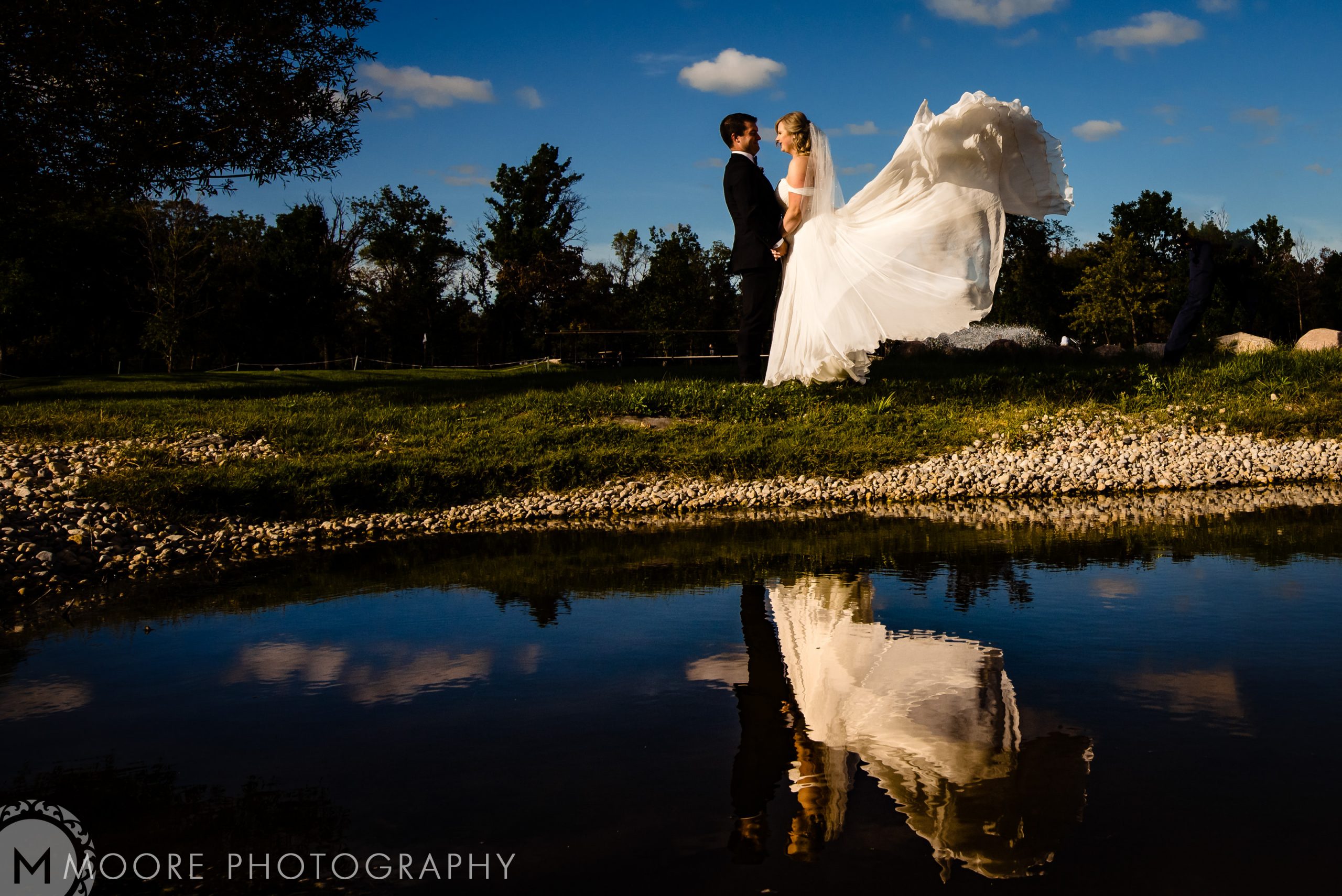 Bride and groom stand mirrored in water under Winnipeg's blue sky with scattered clouds.