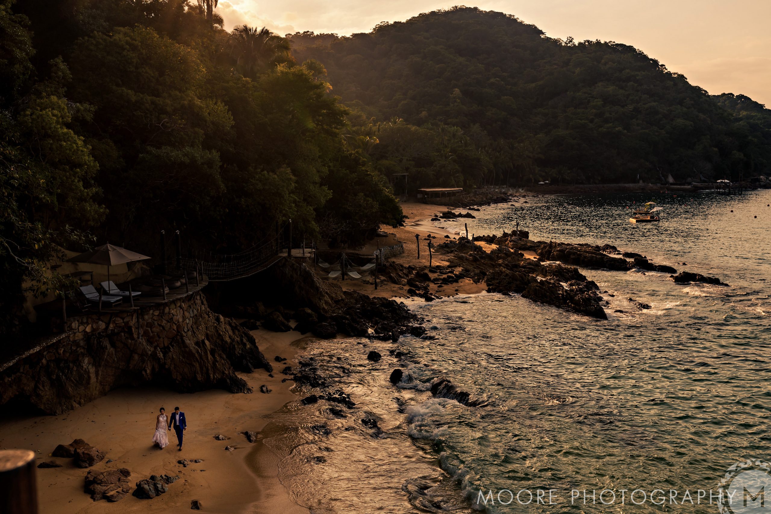 A couple strolls a secluded Puerto Vallarta beach at sunset, framed by trees and rocky shoreline.