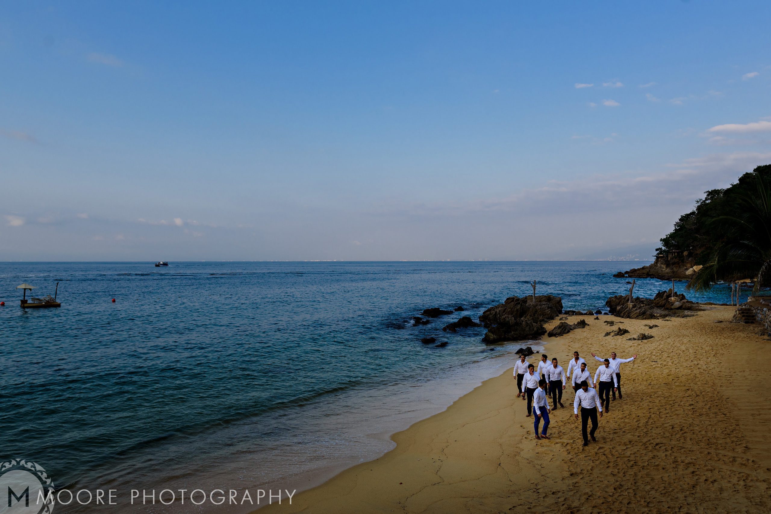 People stroll a Puerto Vallarta beach, rocky outcrops framing a boat under the blue sky.