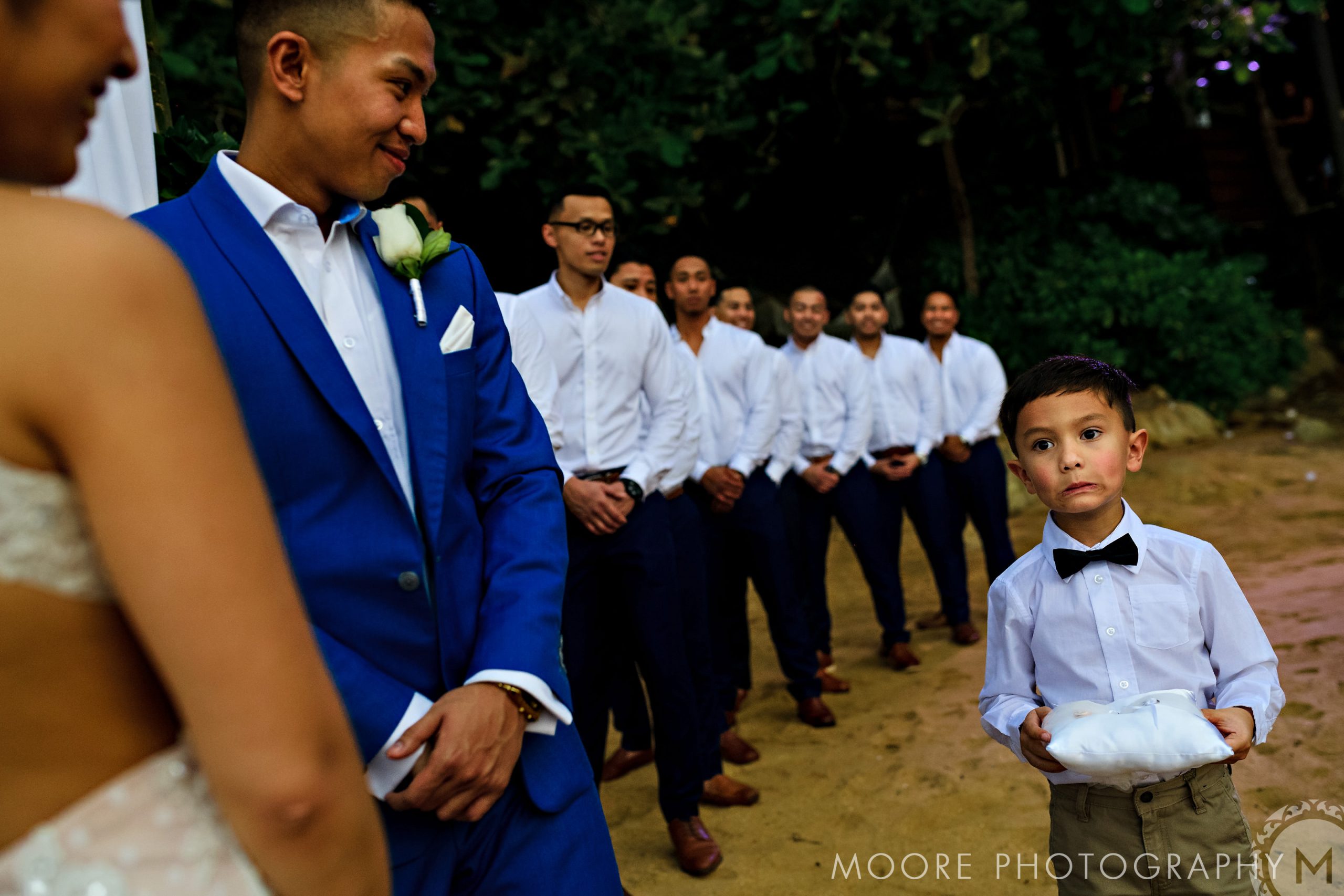Groom in blue suit smiles at bride; young boy holds pillow. Puerto Vallarta Destination Wedding.