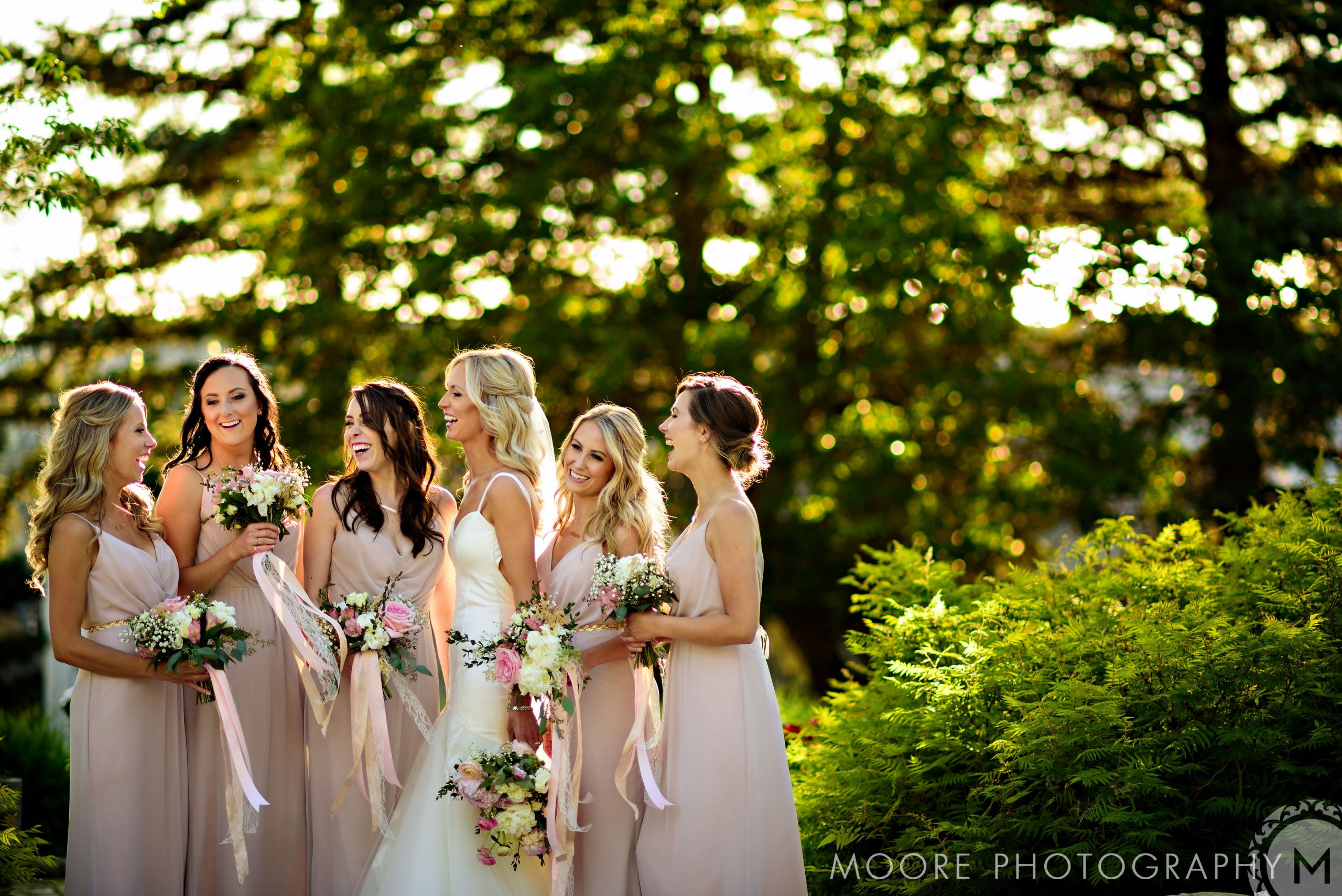 Bridesmaids in blush surround a smiling bride outdoors at a lush Winnipeg wedding venue.