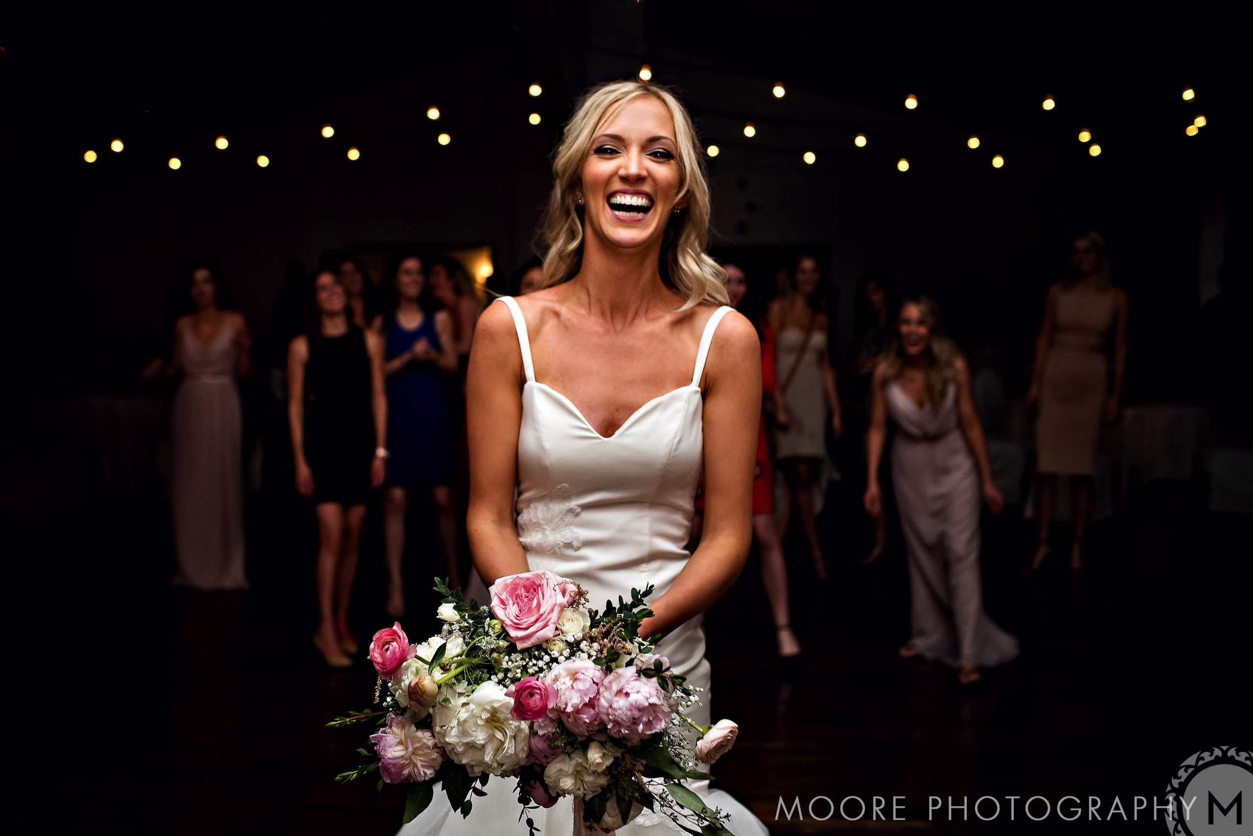 Bride smiling with bouquet, bridesmaids in the background, string lights above—a wedding celebration.