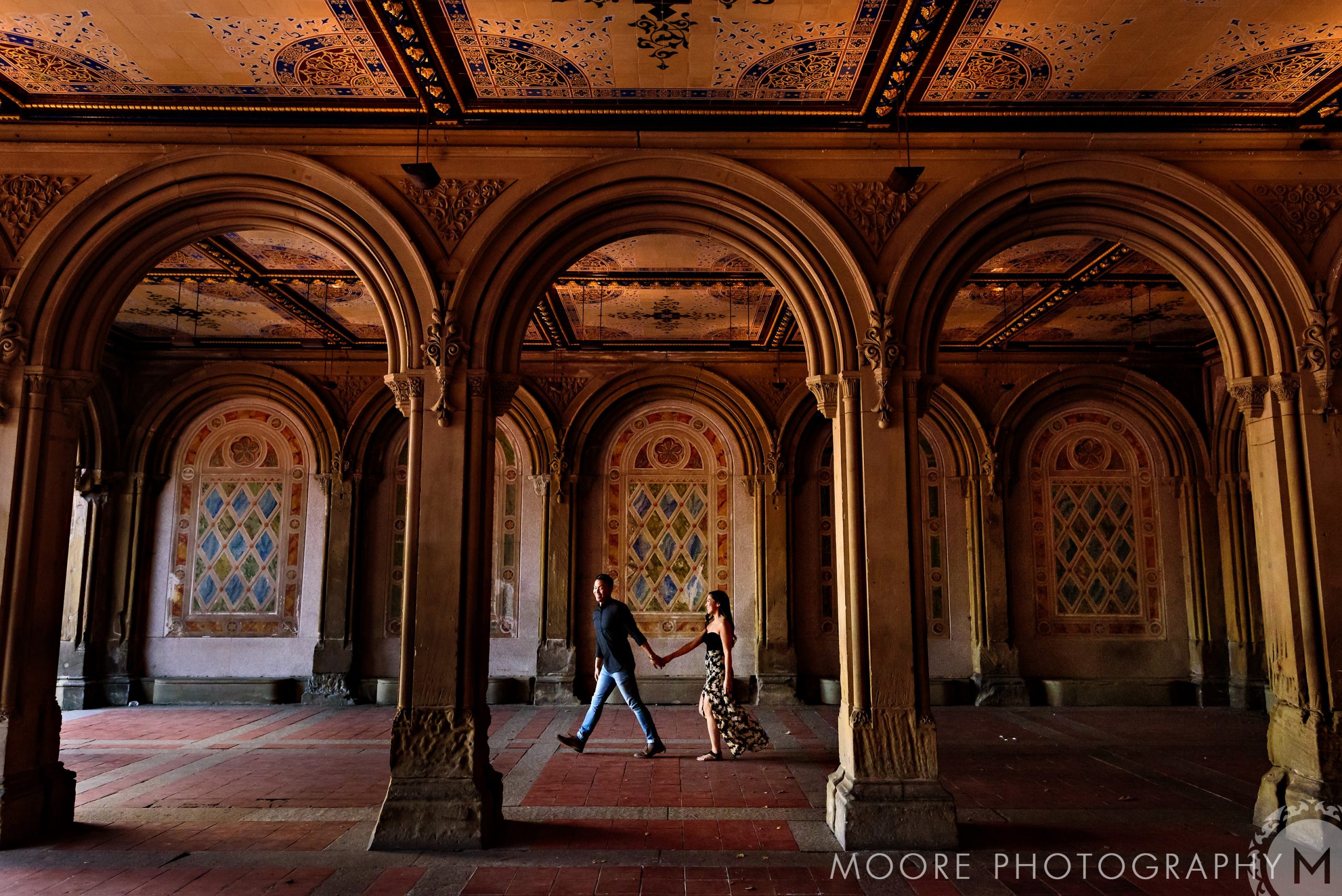 Romantic couple holding hands in ornate, arch-filled hall with decorative ceiling.