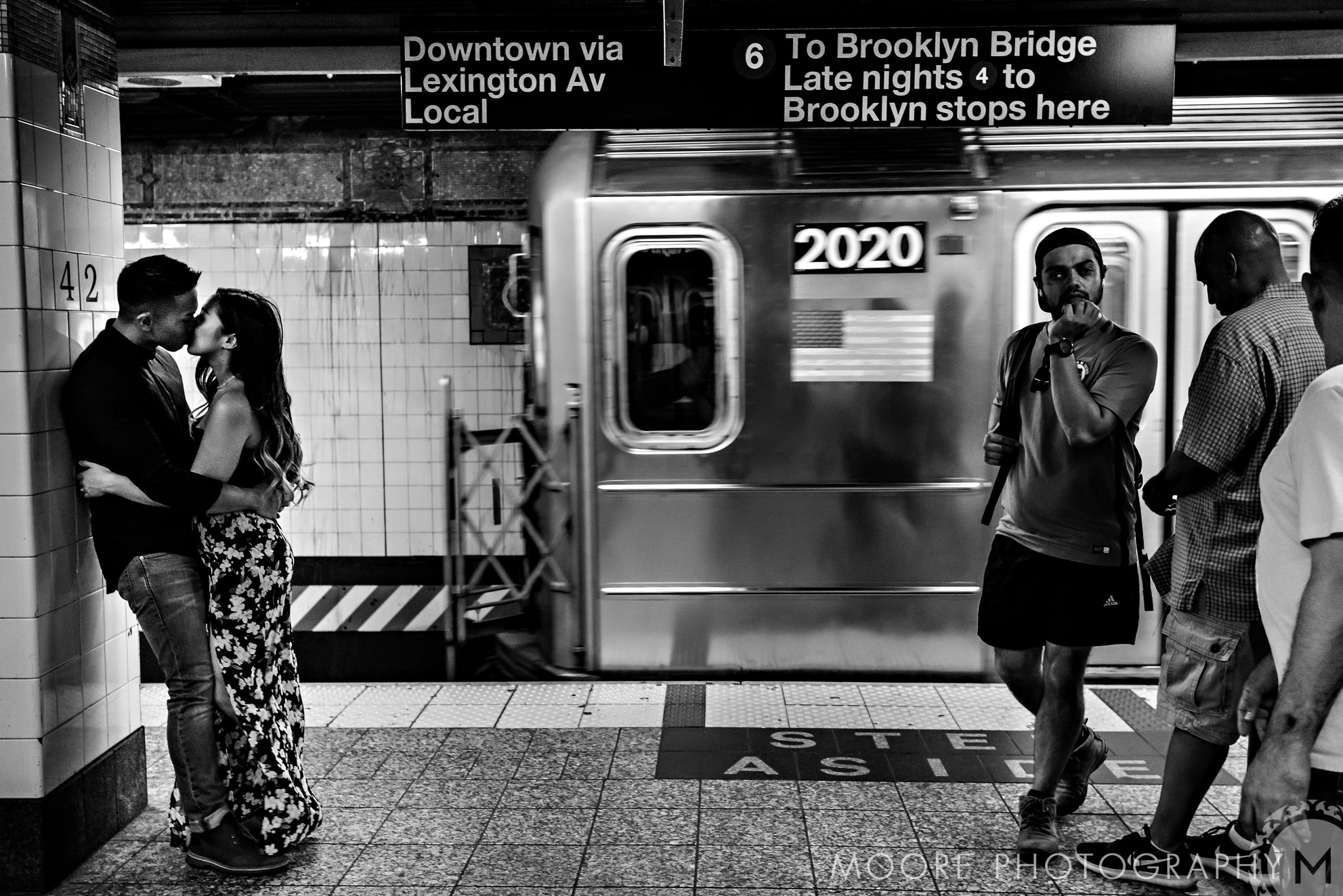 Romantic NYC engagement: couple kissing on subway platform as train passes, people walk by.