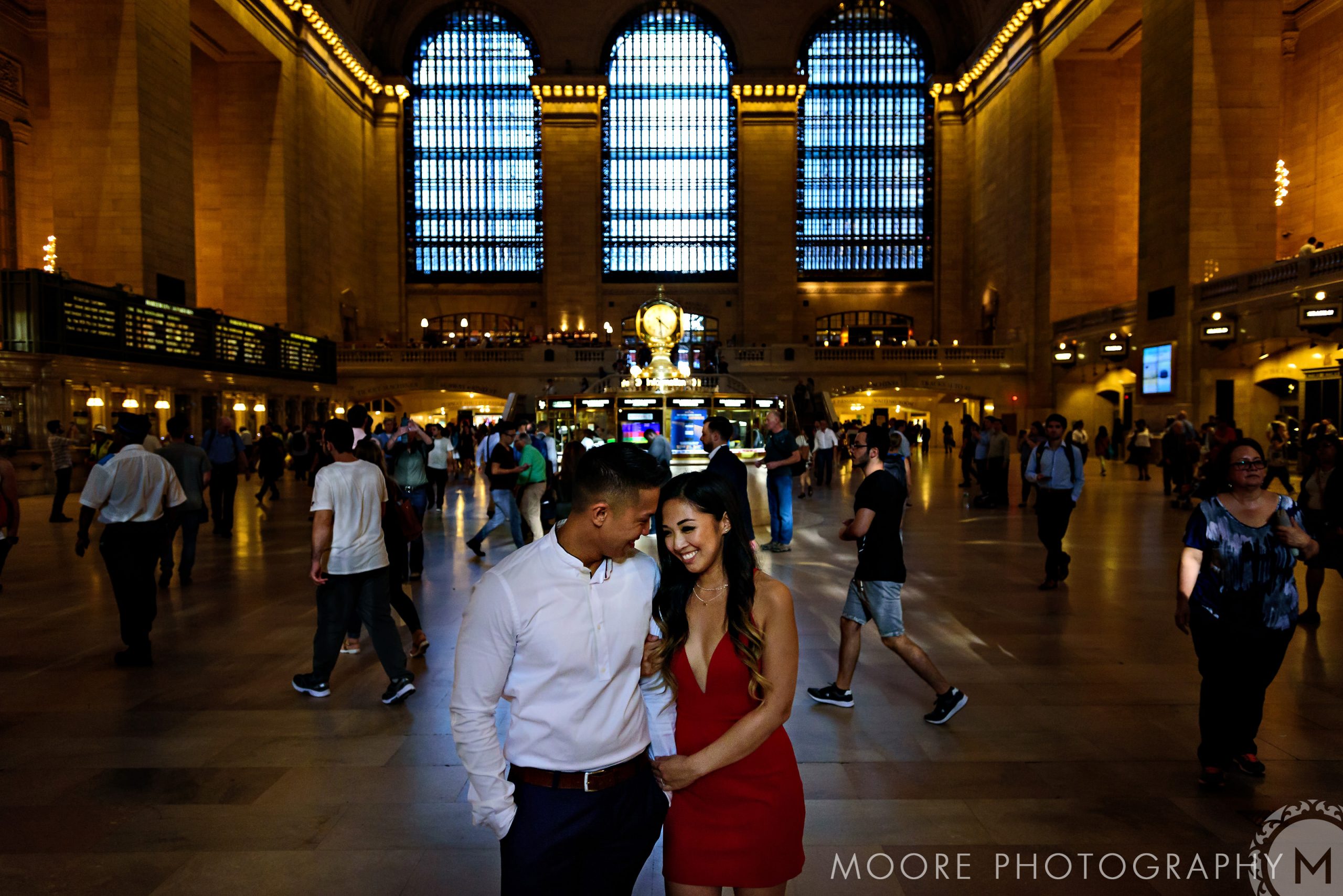 Romantic couple stands together in bustling Grand Central Terminal for NYC engagement photos.