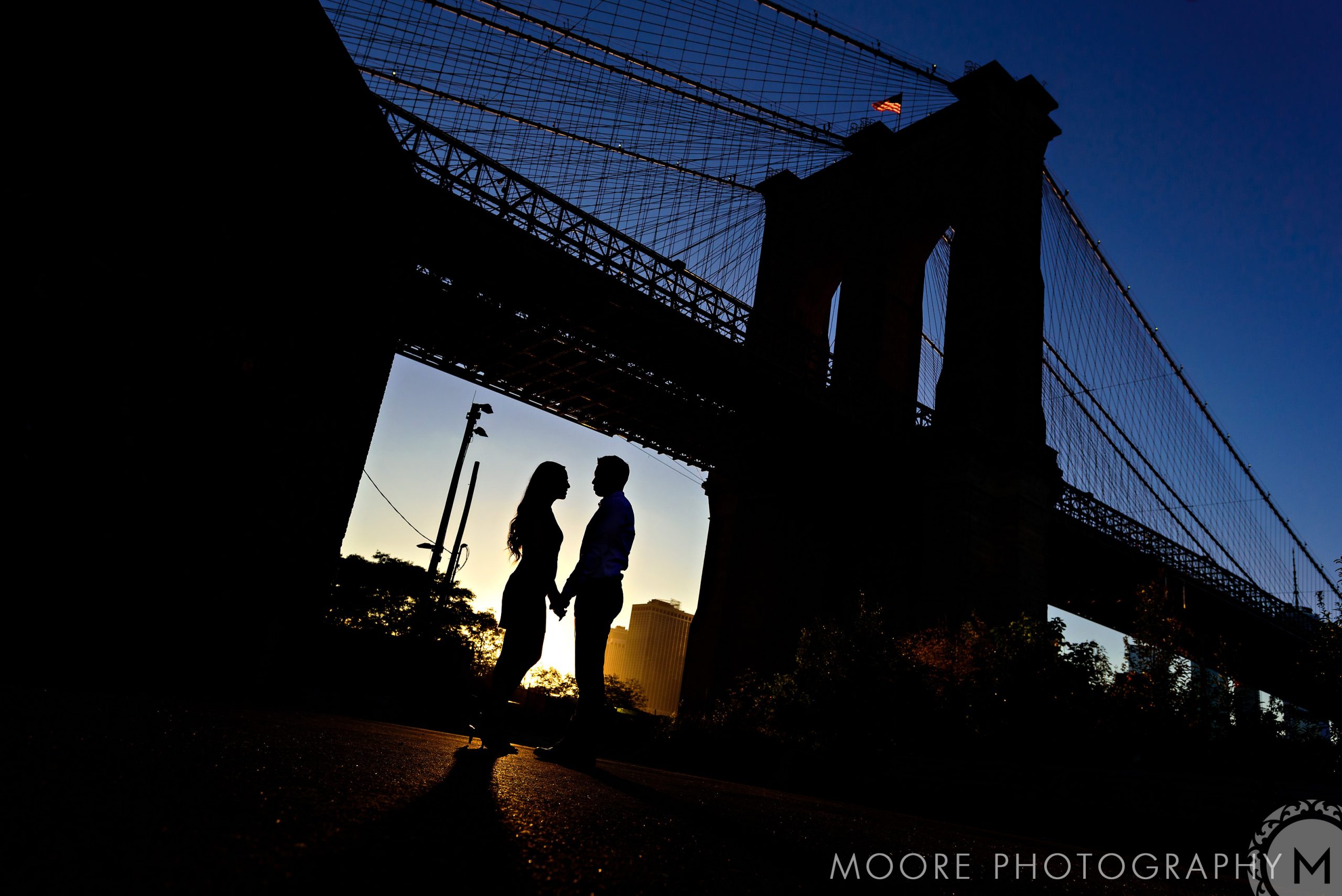 Romantic couple holding hands under a NYC bridge at sunset.