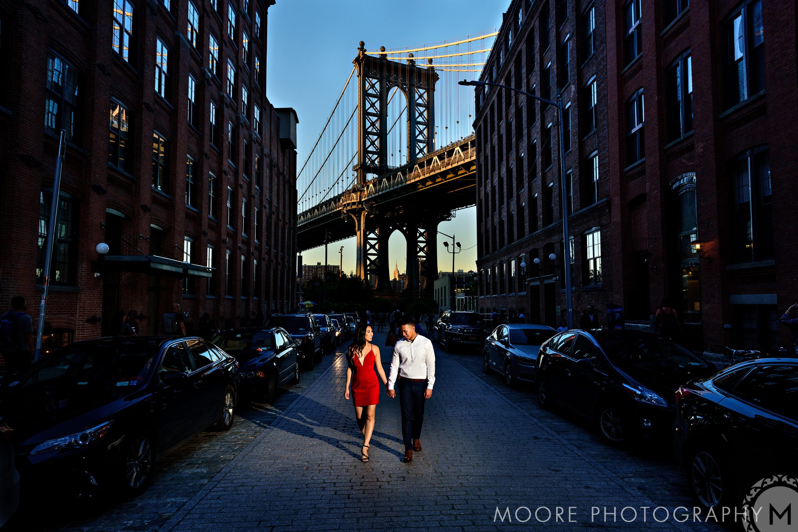 Romantic couple walking a cobblestone street with an NYC bridge backdrop.
