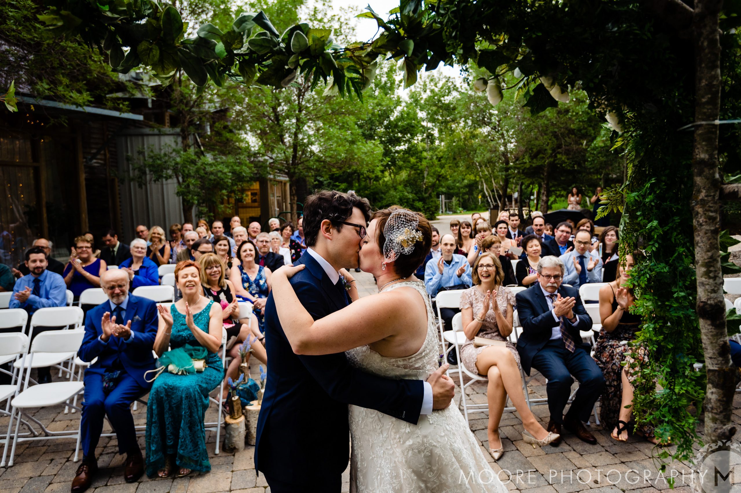 Bride and groom kiss at outdoor Winnipeg wedding, guests applauding behind them.