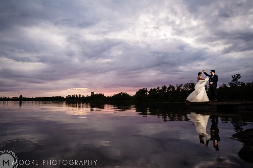 I bride and groom, dancing for a wedding photo with a lake and sunset in the background at Fort Whyte in Winnipeg
