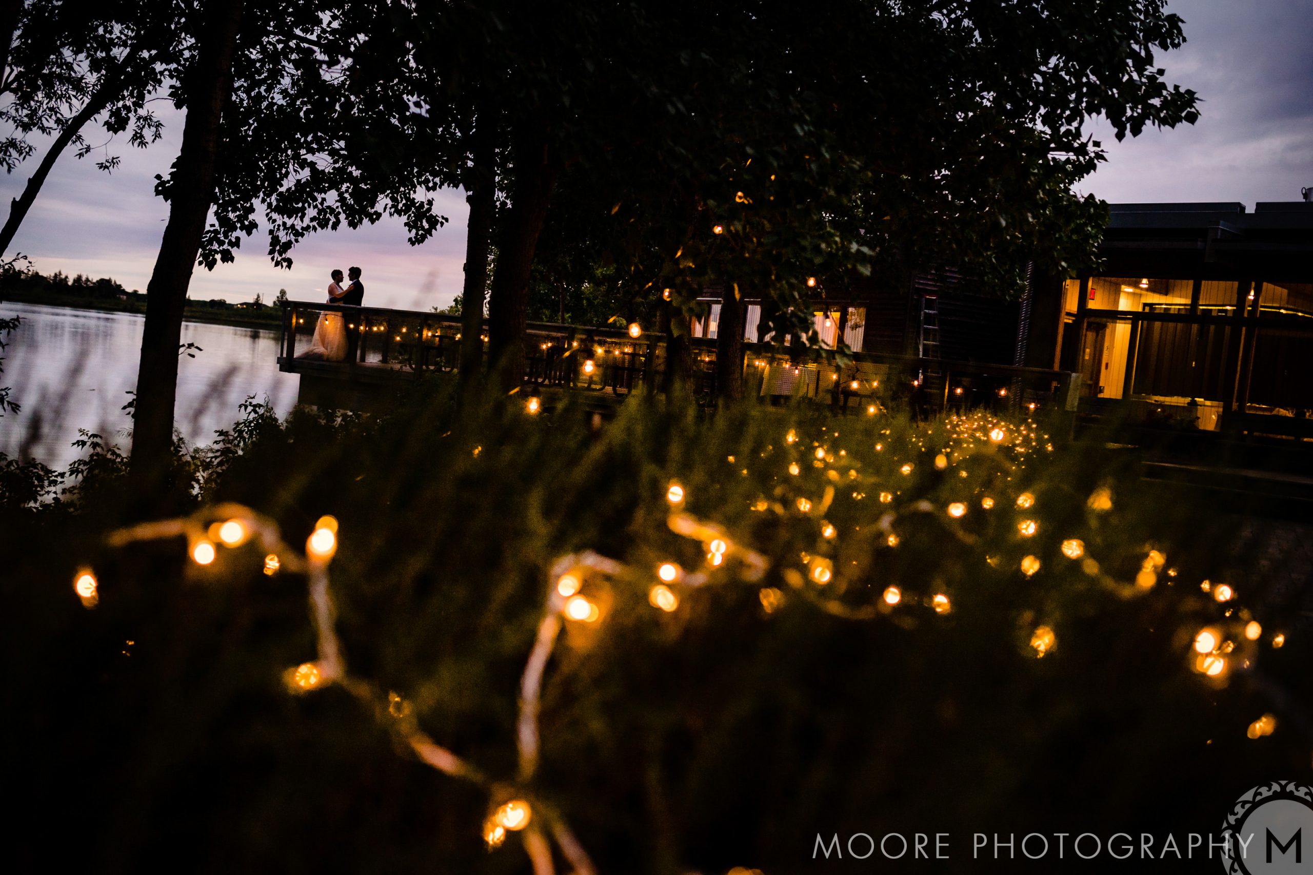 Couple at dusk by a lake, like Winnipeg wedding venues with warm lights on bushes.