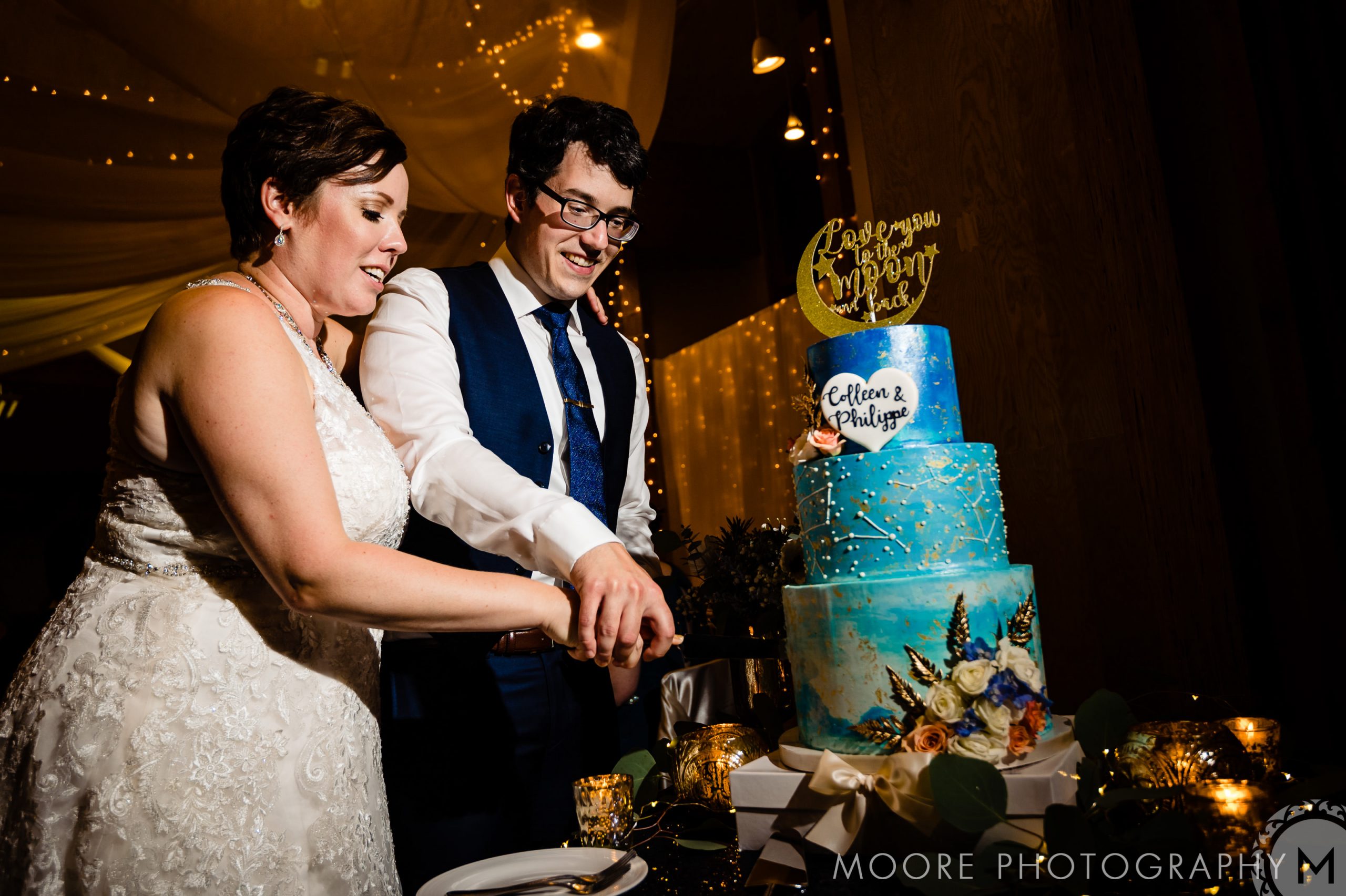 Couple slicing a blue wedding cake under string lights at a Winnipeg wedding venue.