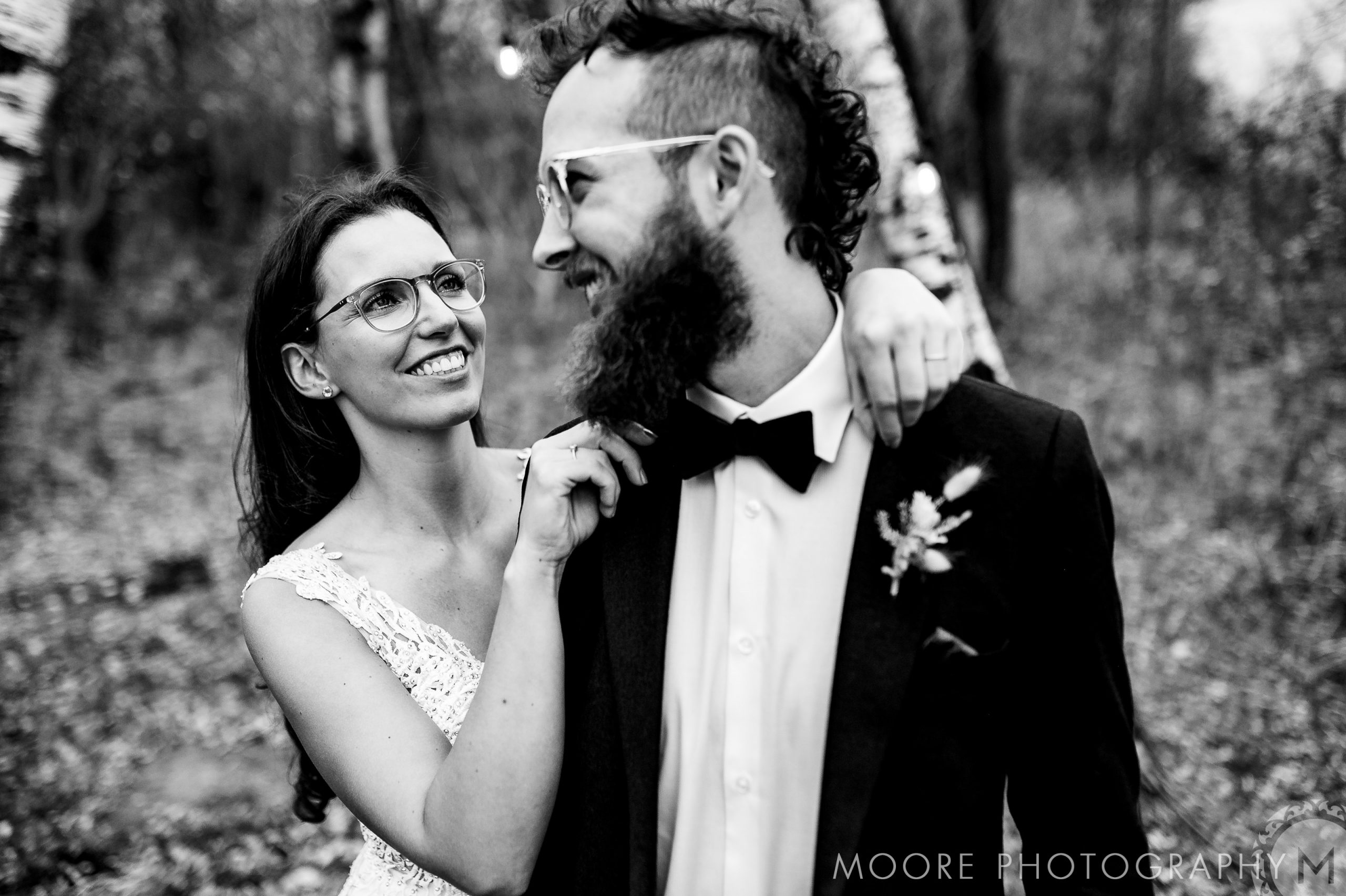 Bride adjusts groom's bow tie, both smiling in a forest. Winnipeg wedding venue. Black and white photo.